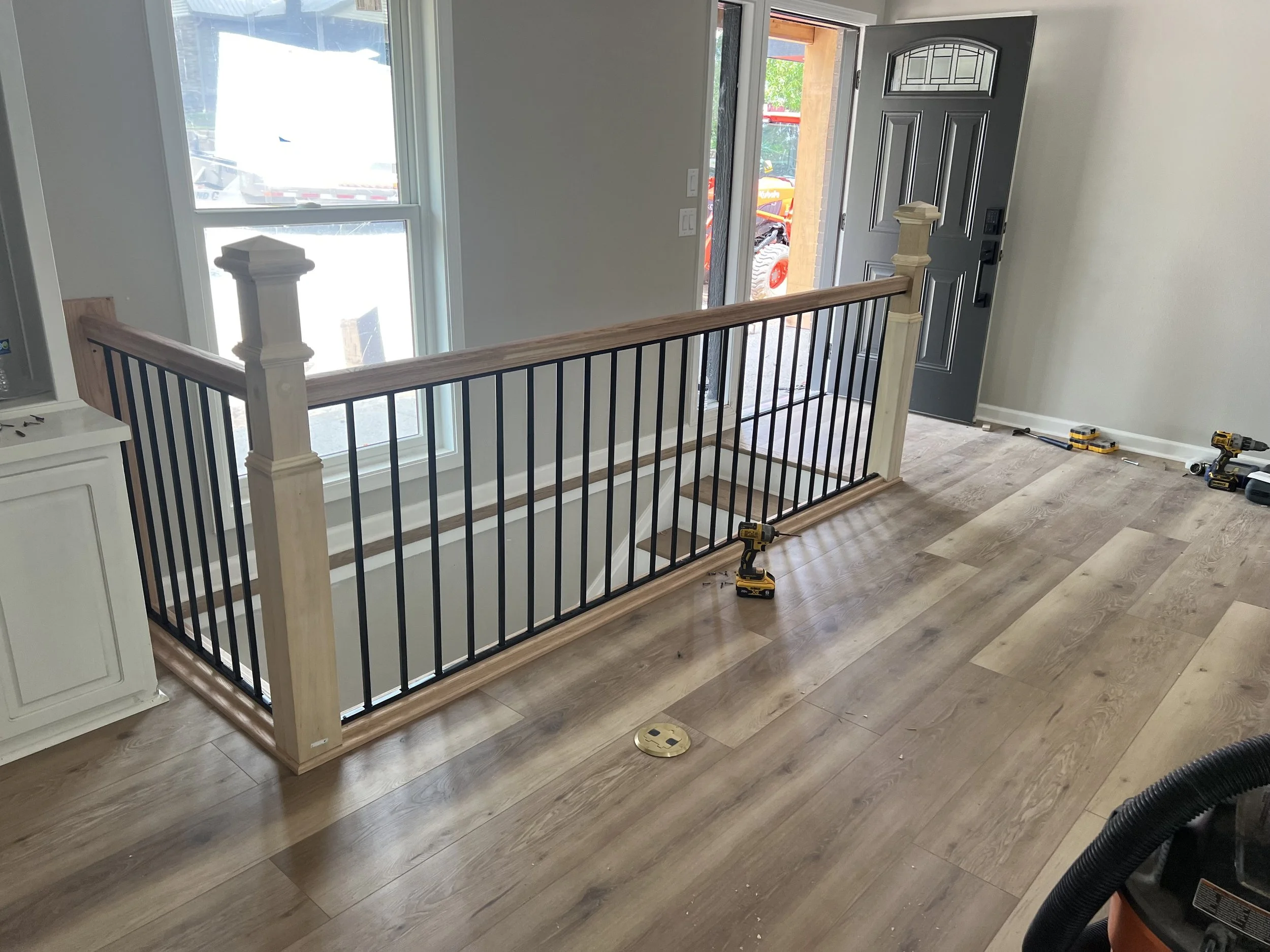 Interior of a house under renovation with newly installed staircase railing, tools on floor, and an open front door leading outside.