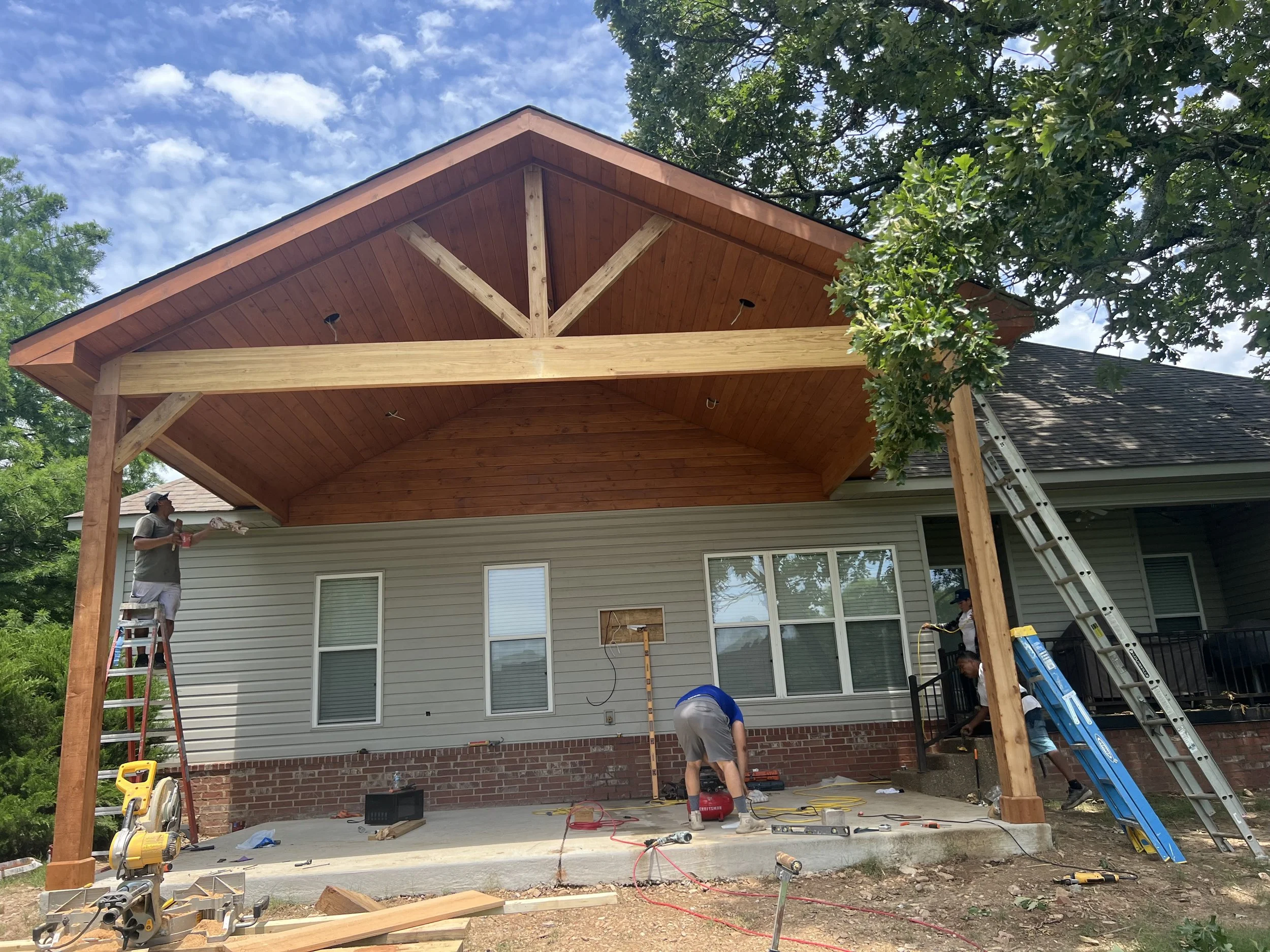 Construction workers building a wooden porch or extension on the back of a house, with ladders, tools, and construction materials visible.
