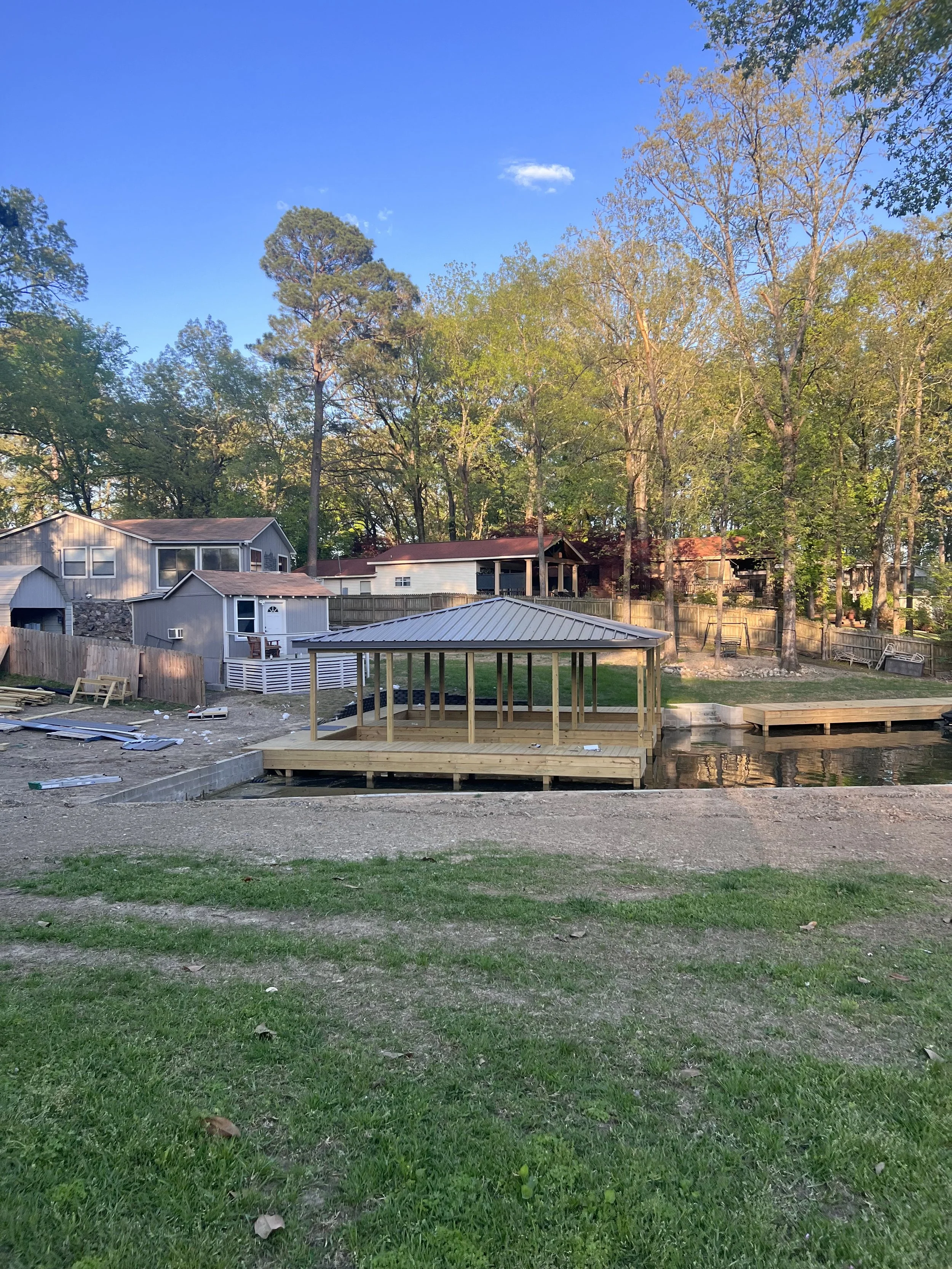 Residential backyard with green lawn, wooden deck under construction, surrounding trees, neighboring houses, clear blue sky.