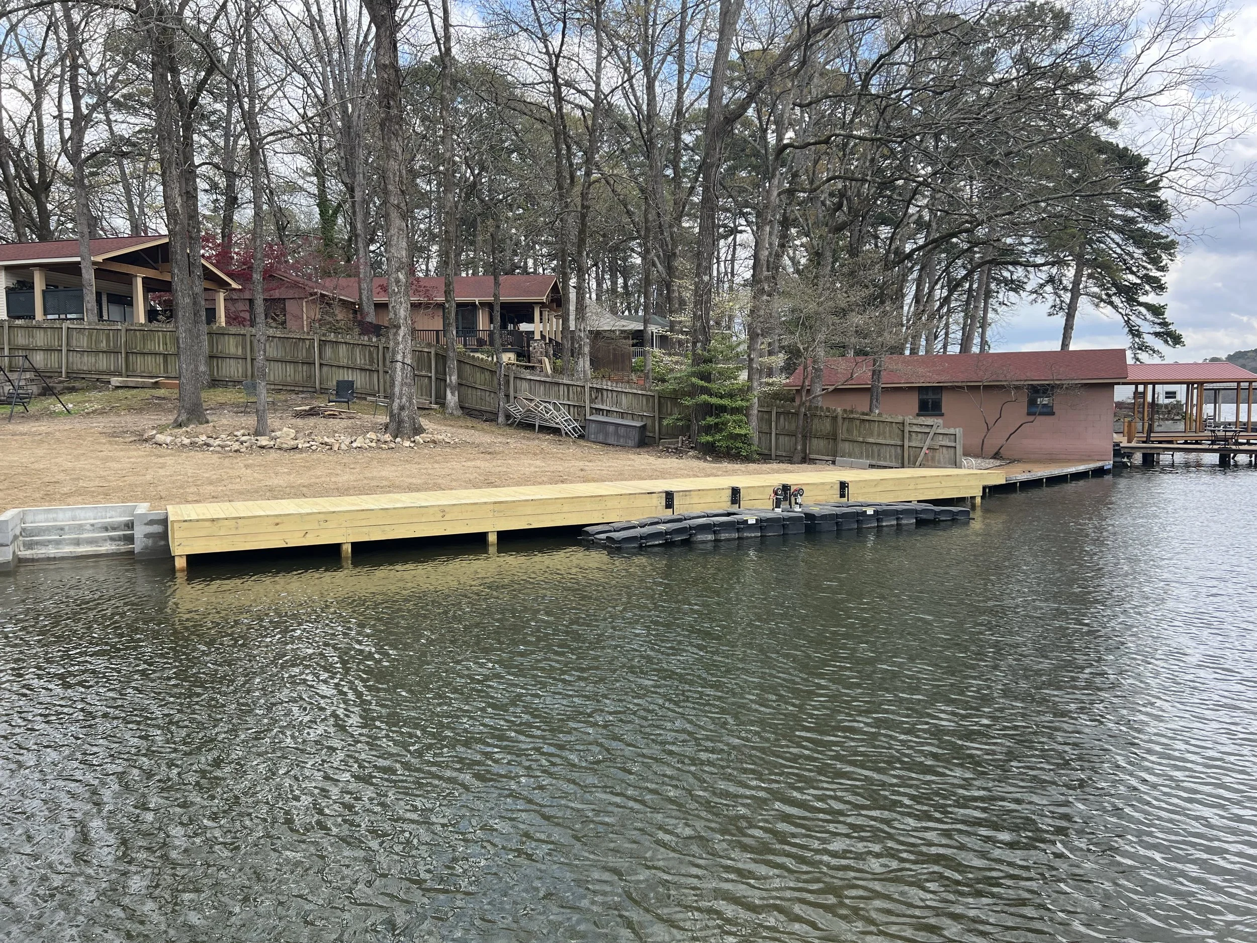 Residential houses by a body of water with a wooden dock, trees with some leaves, and a cloudy sky.