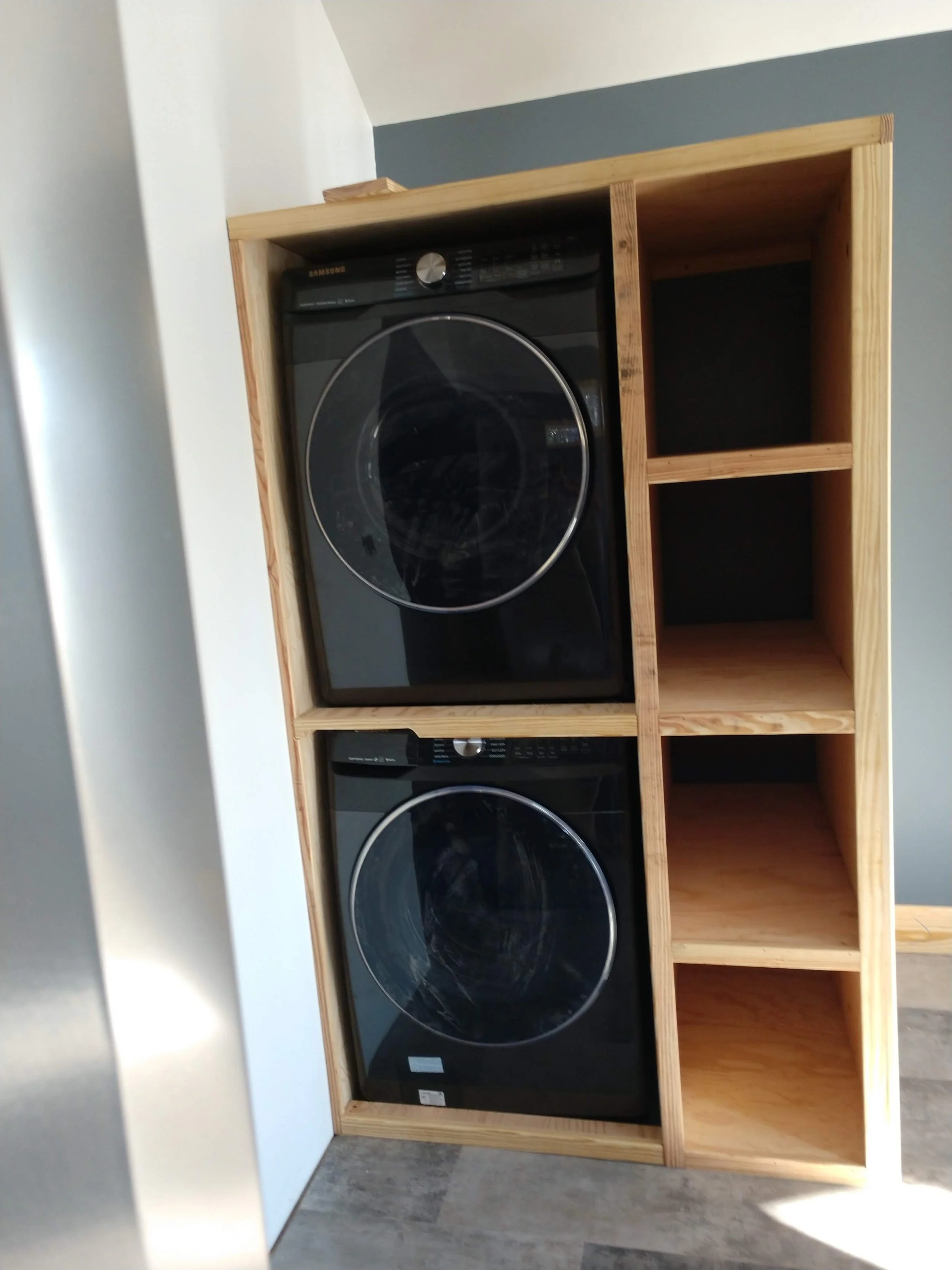 A black washing machine with a glass door, placed inside a wooden frame with open compartments to the right and below.