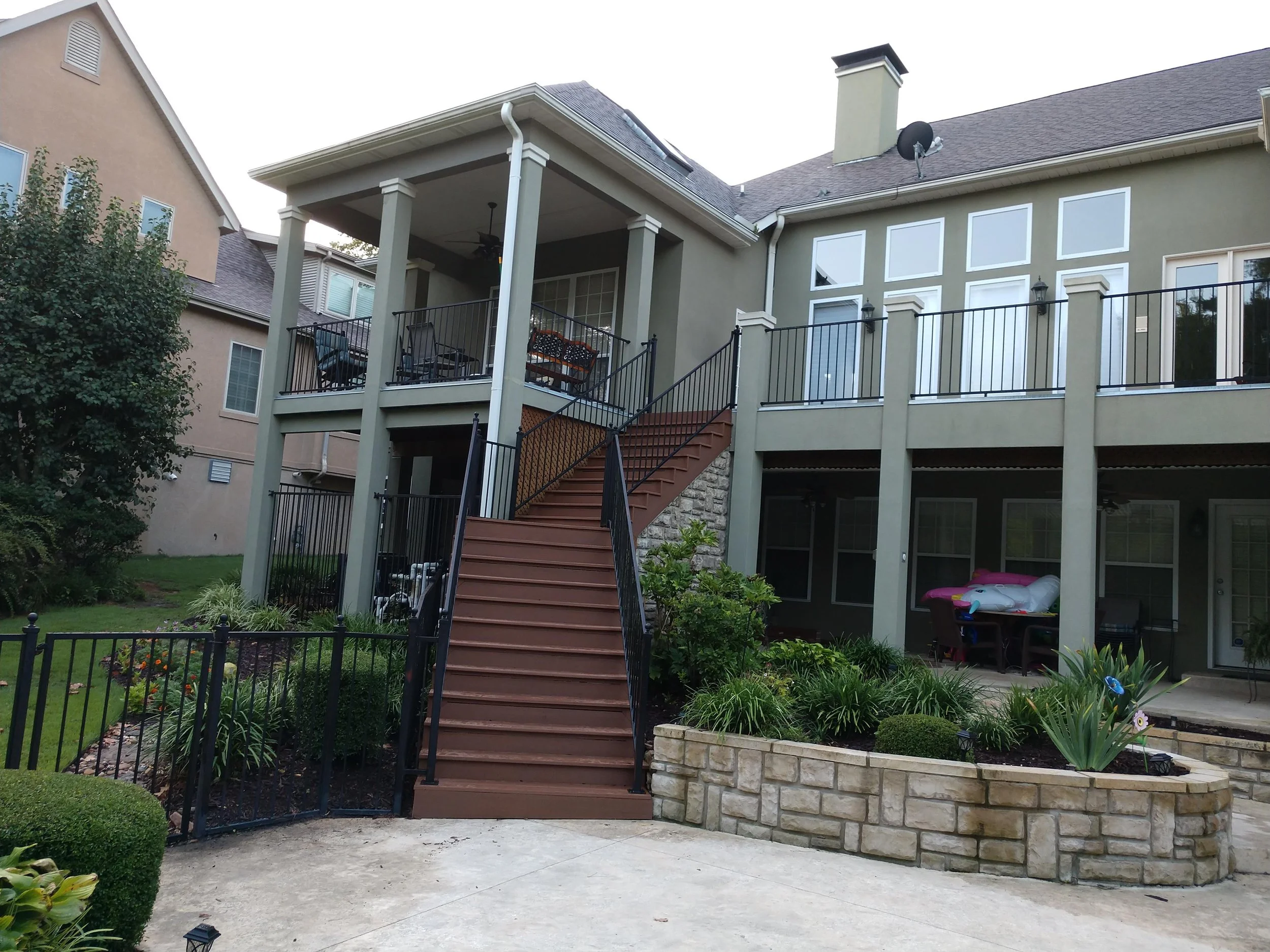 Backyard view with a raised deck and stairs, surrounded by a well-manicured garden and lawn, with neighboring houses visible in the background.