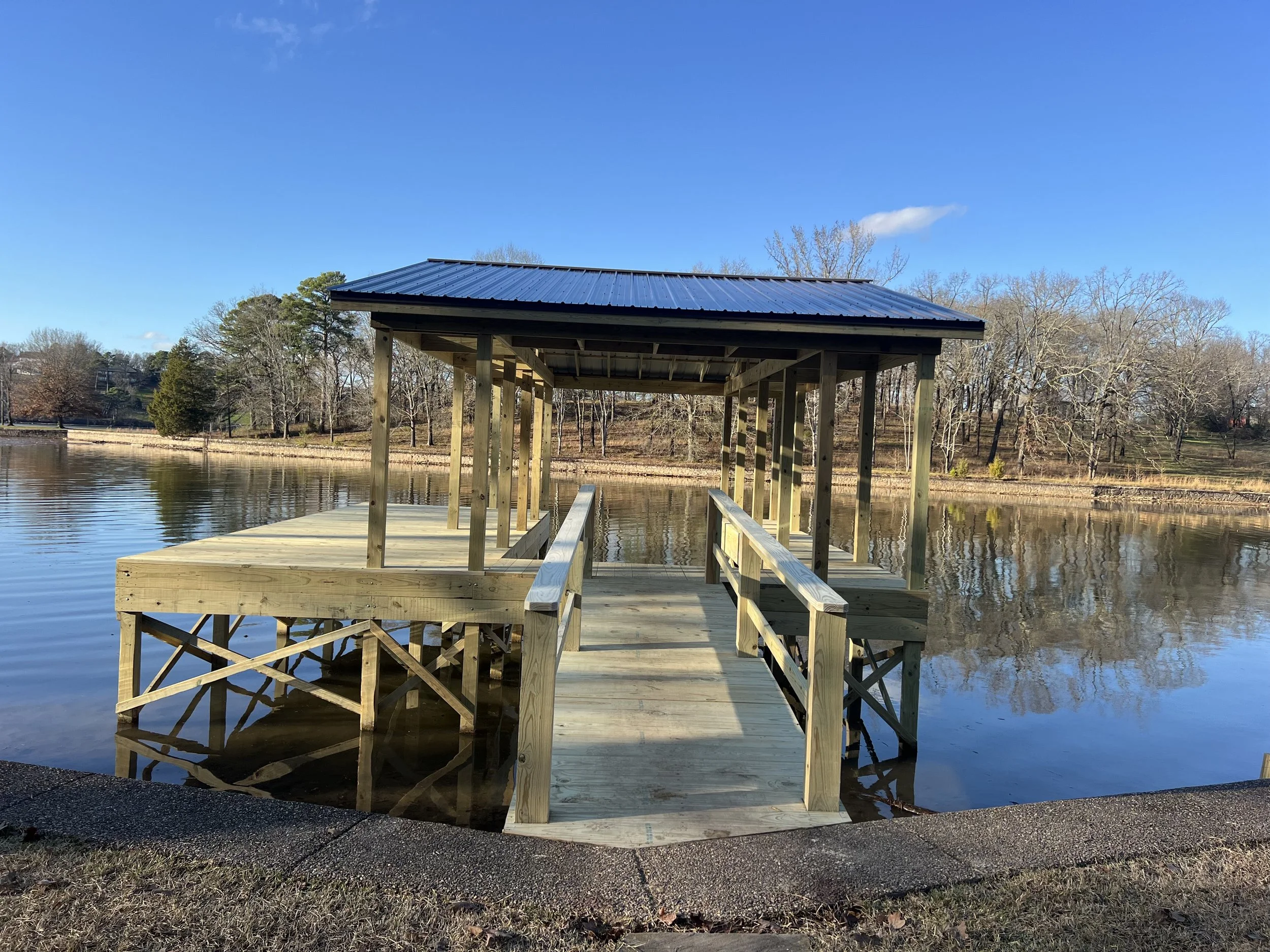 A wooden dock with a small covered fishing pier extends over a calm lake with trees in the background under a clear blue sky.