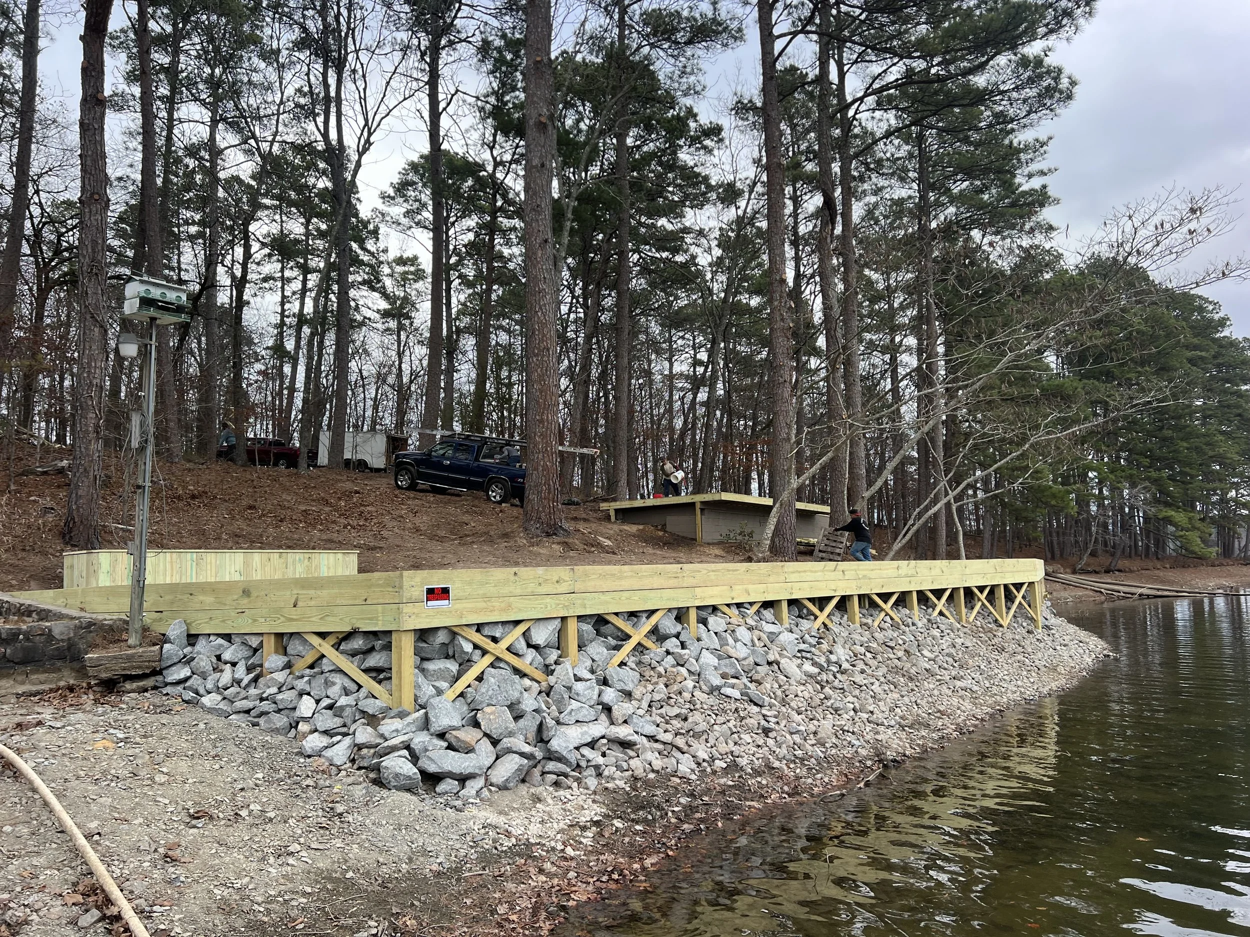 A lakeside construction site with a wooden deck supported by stone-filled gabion walls, surrounded by tall pine trees and parked vehicles, with an overcast sky.