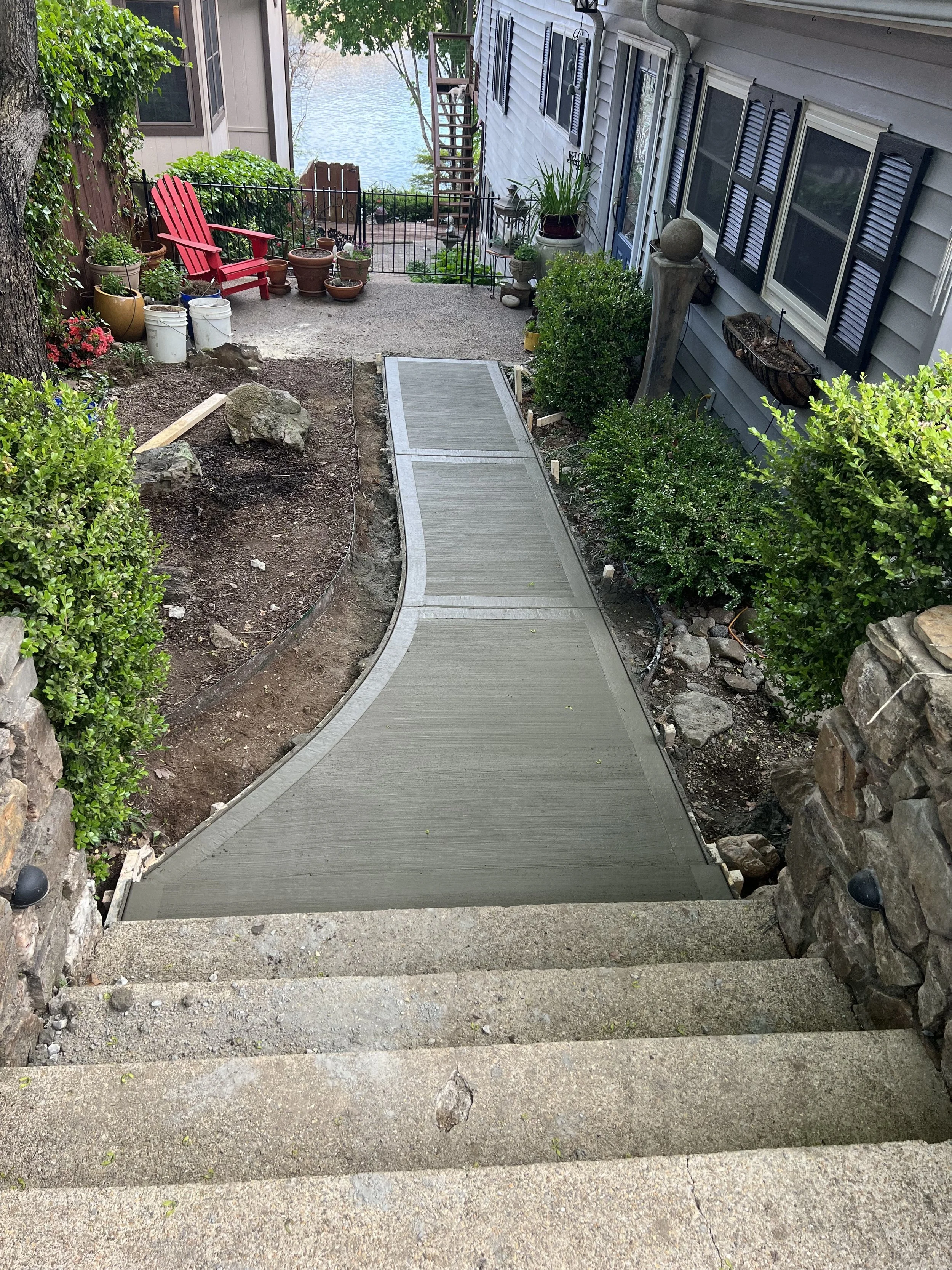 A view looking down steps onto a paved pathway in a backyard. The pathway leads to a small patio area with a red Adirondack chair, various potted plants, and a lake in the background. The house's exterior has light-colored siding and several windows 