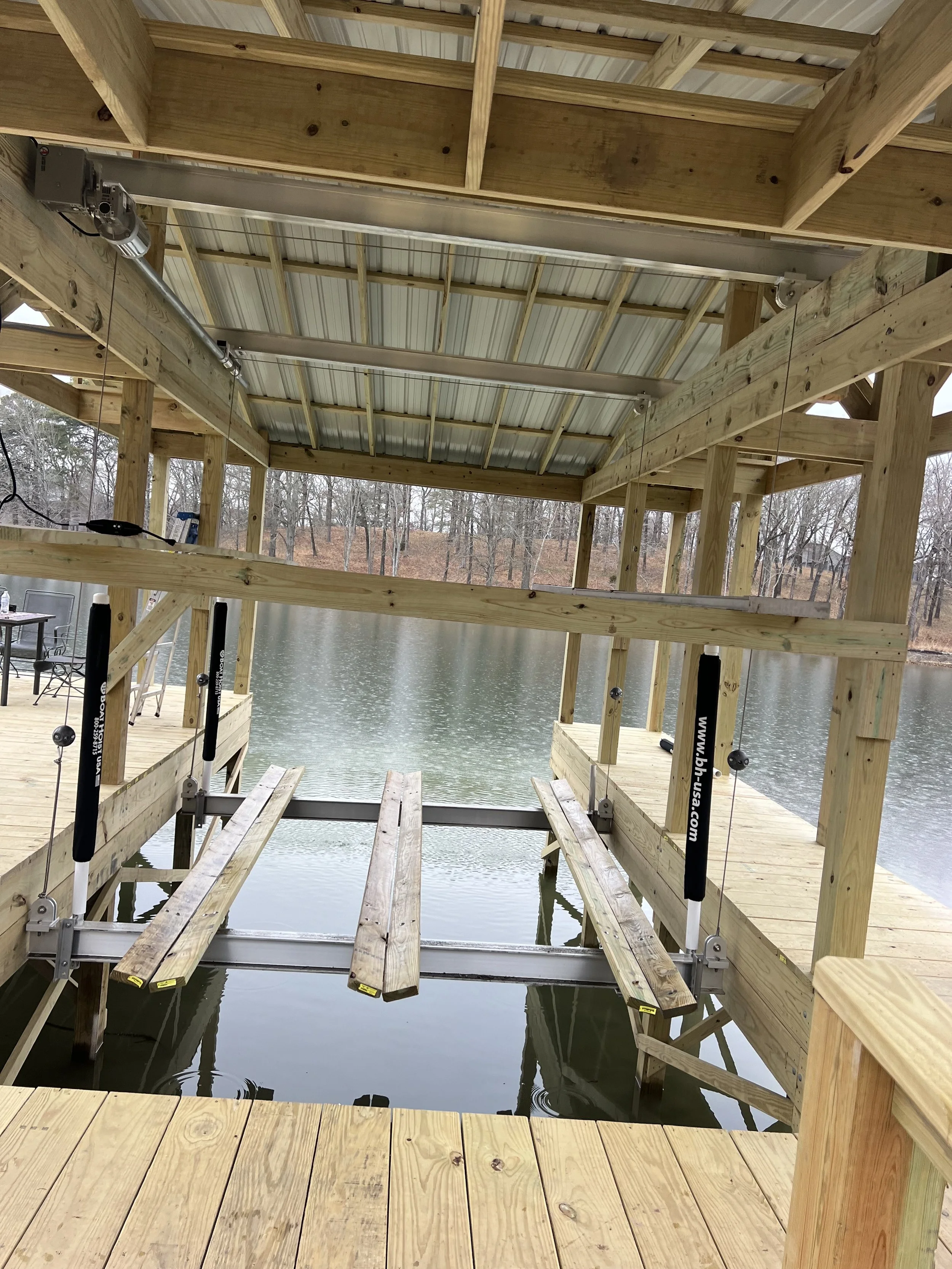 Wooden dock extending over a body of water, with a partially constructed wooden structure on top, supported by vertical beams, and metal framing and tools visible.