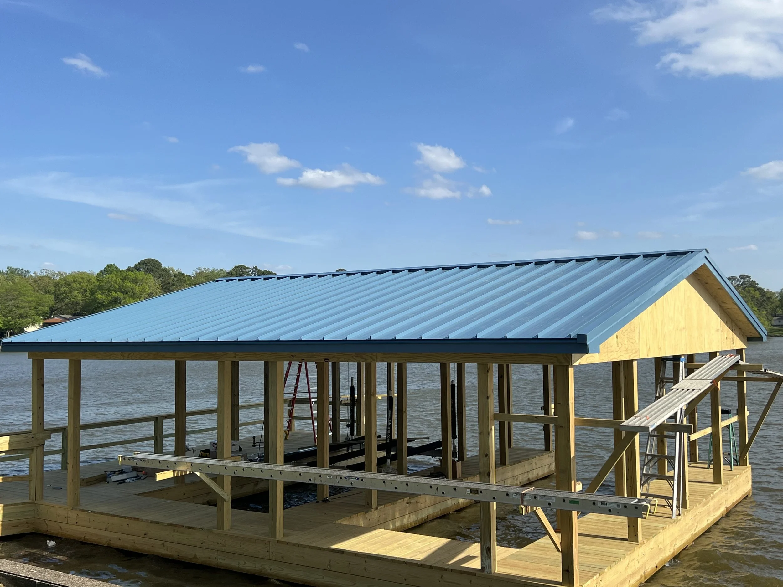 A wooden dock under construction over a body of water with a building that has a blue metal roof and yellow walls. Trees are in the background, and the sky is blue with scattered white clouds.