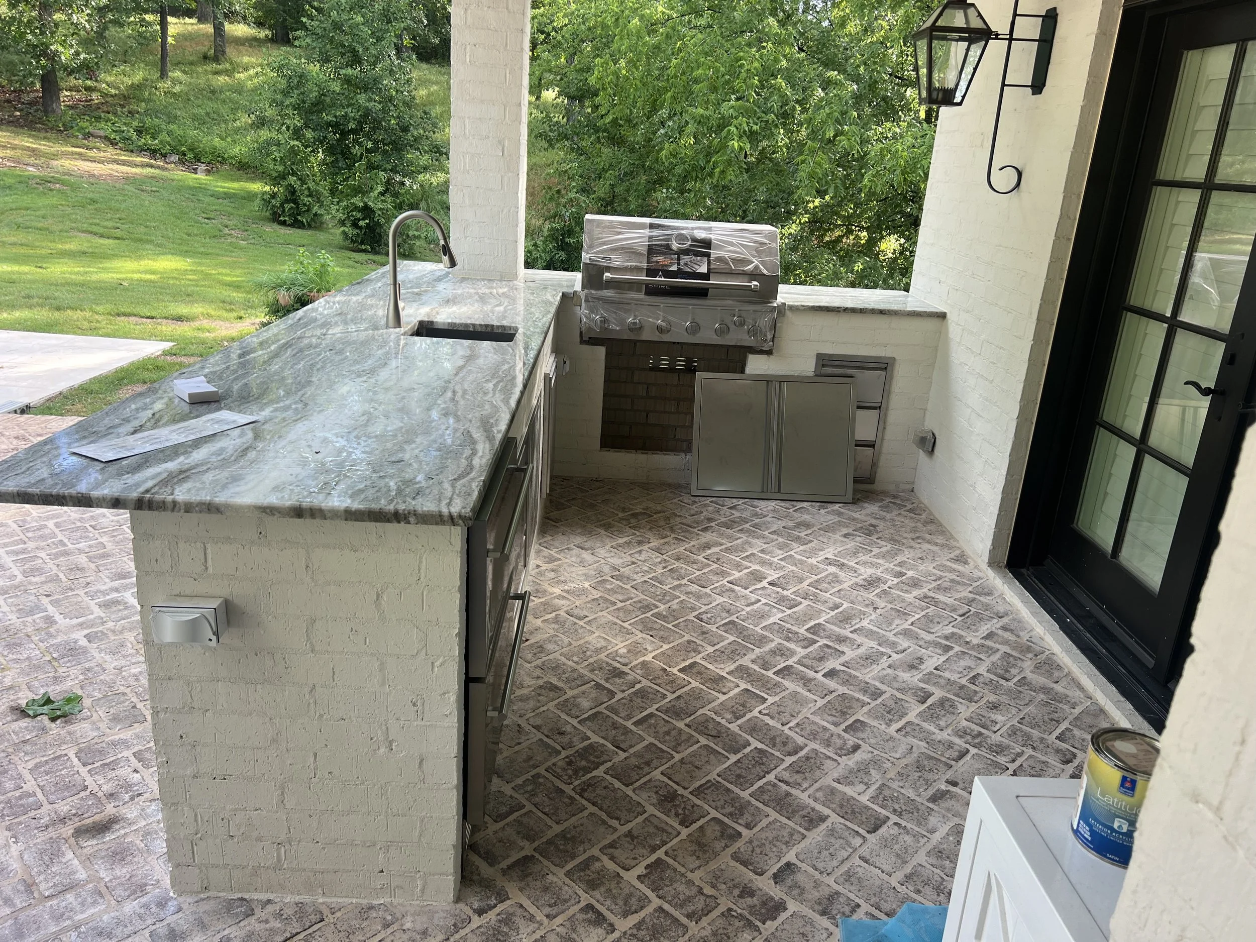 An outdoor kitchen area with a marble countertop, a stainless steel grill covered with plastic, a sink with a modern faucet, and a view of green trees and grass in the background. There is also a black lantern-style light fixture on the wall and a sm