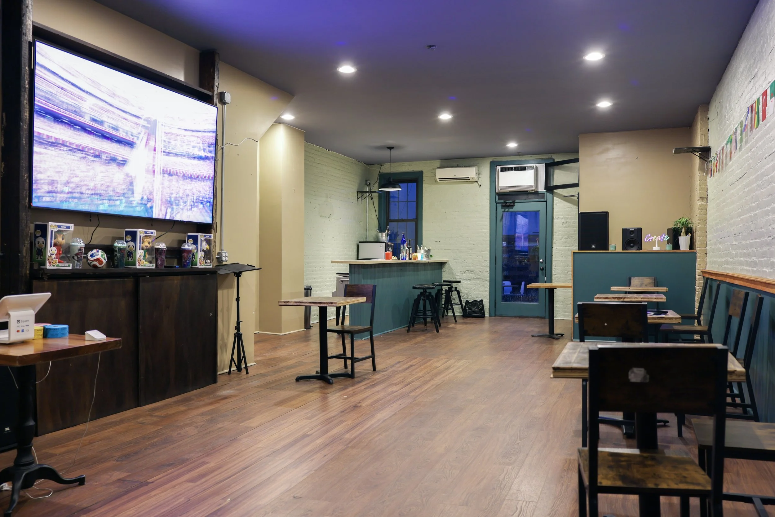 Interior of a cozy restaurant or cafe with wooden flooring, several tables and chairs, a large flat-screen TV mounted on the wall, and decorations including colorful banners and plants. There are large windows and a door at the back, with air conditioning units visible on the walls.