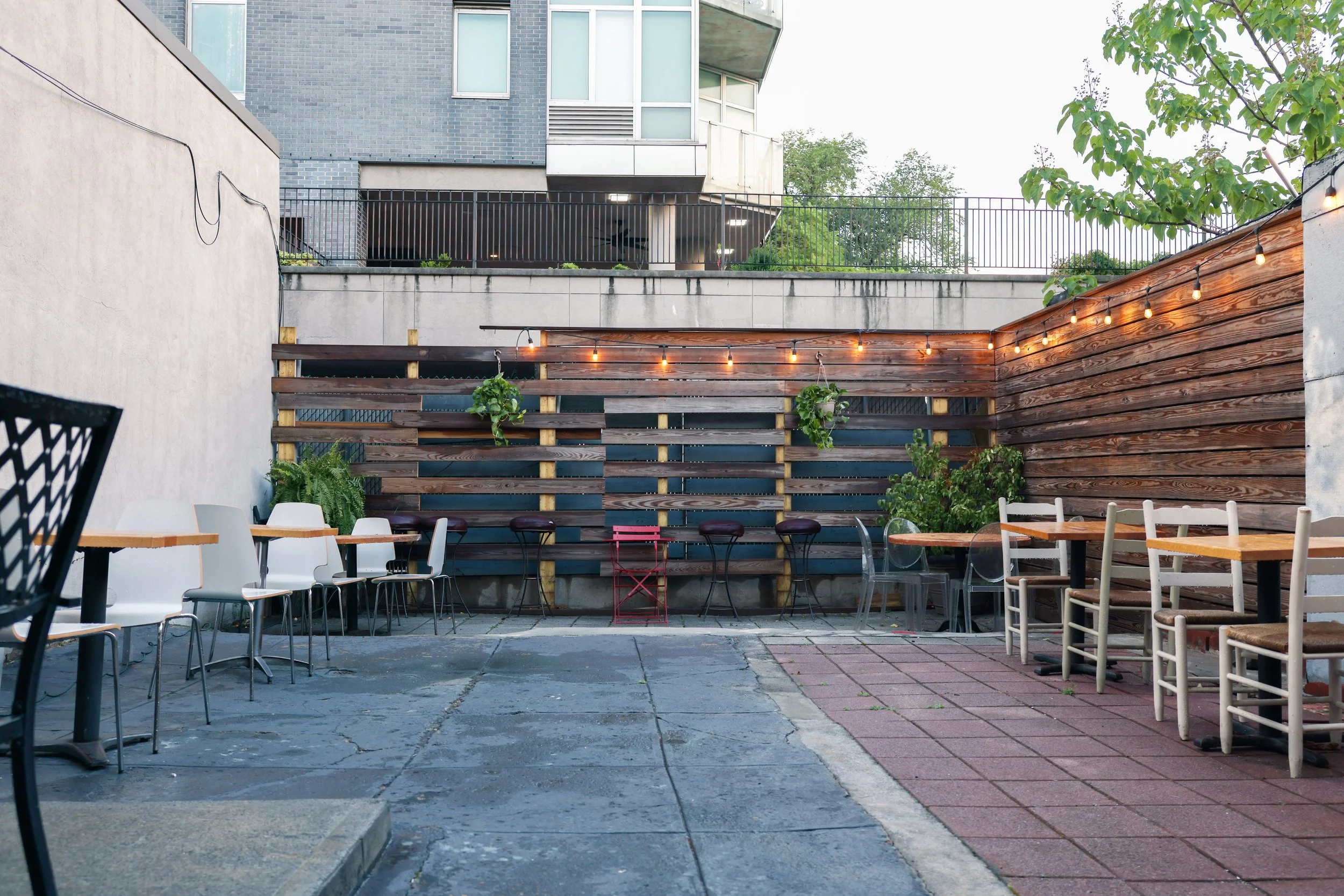 An outdoor patio area with chairs and tables, surrounded by a wooden fence with hanging string lights. There are some plants and a tree, with a modern multi-story building in the background.