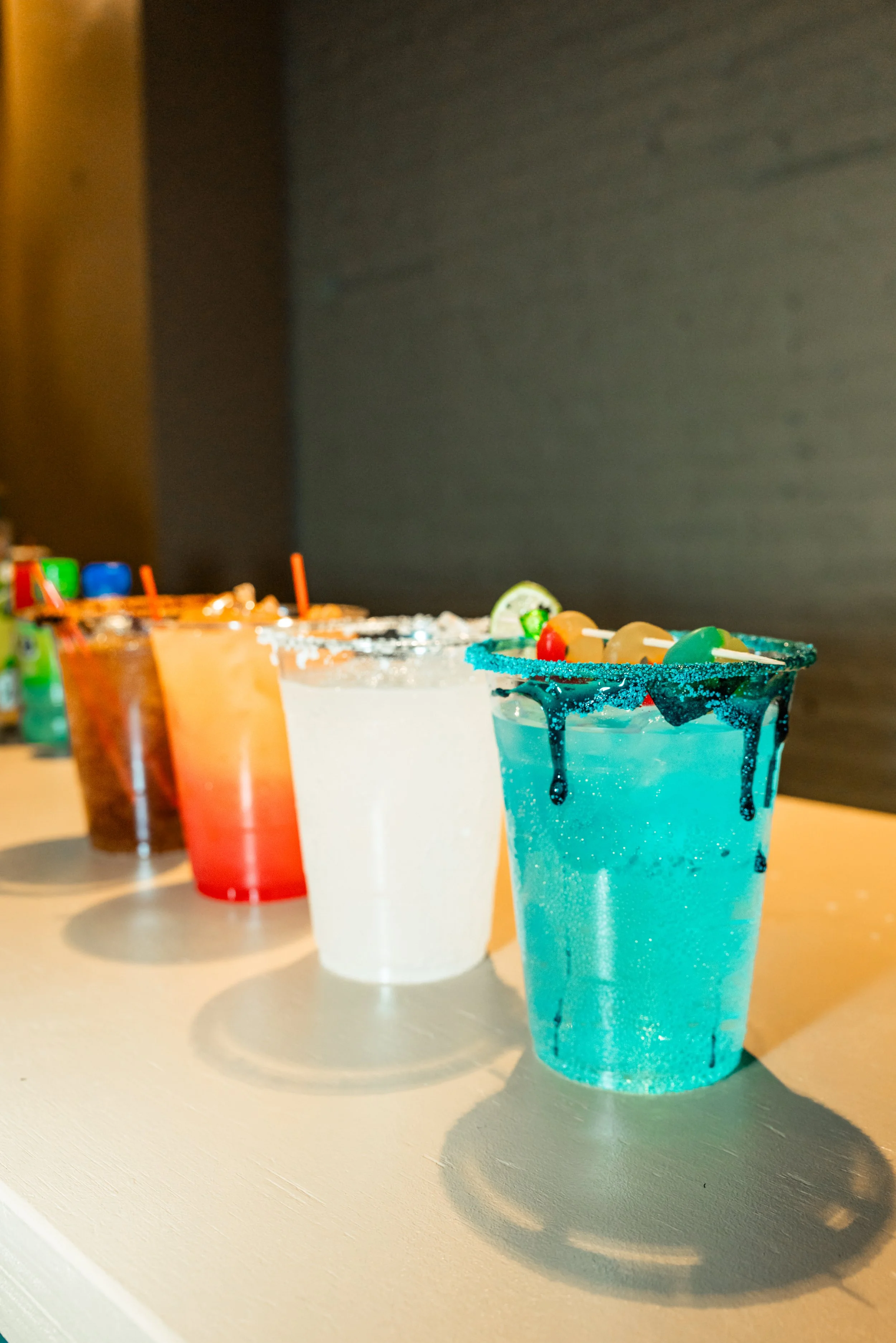 Colorful margarita cocktails with salt and sugar rims, garnished with slices of lime and fruit, lined up on a table.