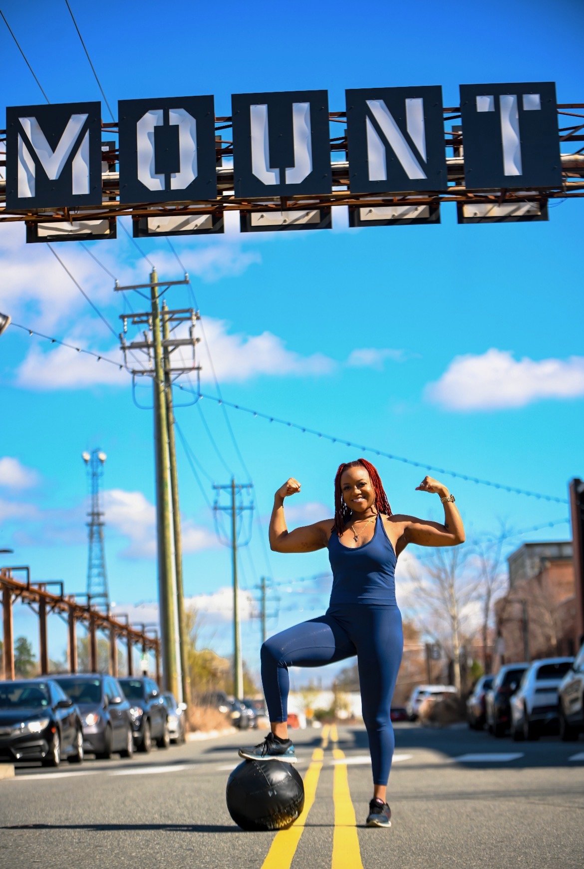 A woman wearing blue workout clothes standing on a yellow-lined street, flexing her biceps while balancing on a large black exercise ball with a weathered building and a clear blue sky in the background.