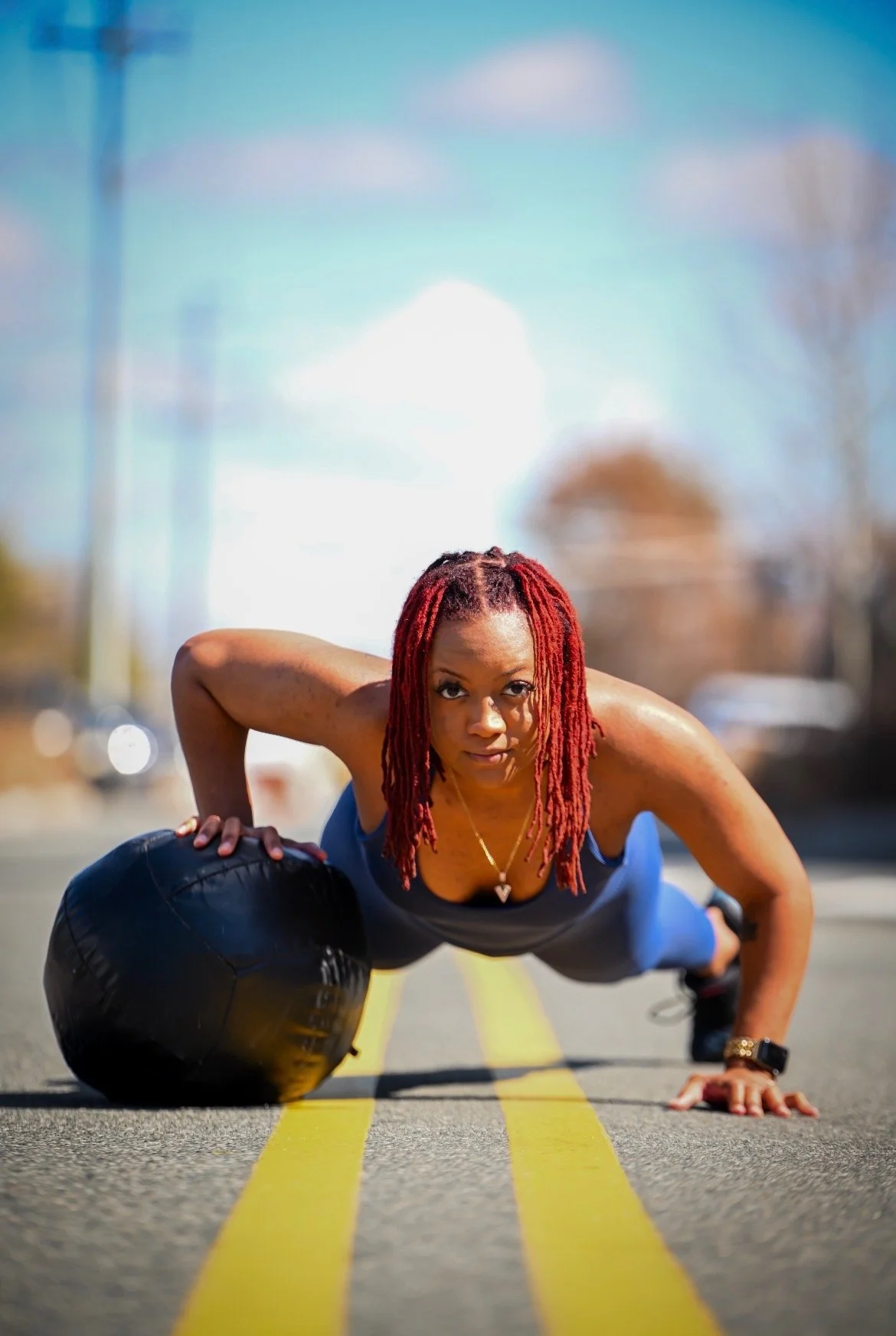 A woman doing a plank exercise on a street, using a large black medicine ball for support, in bright daylight under a blue sky.