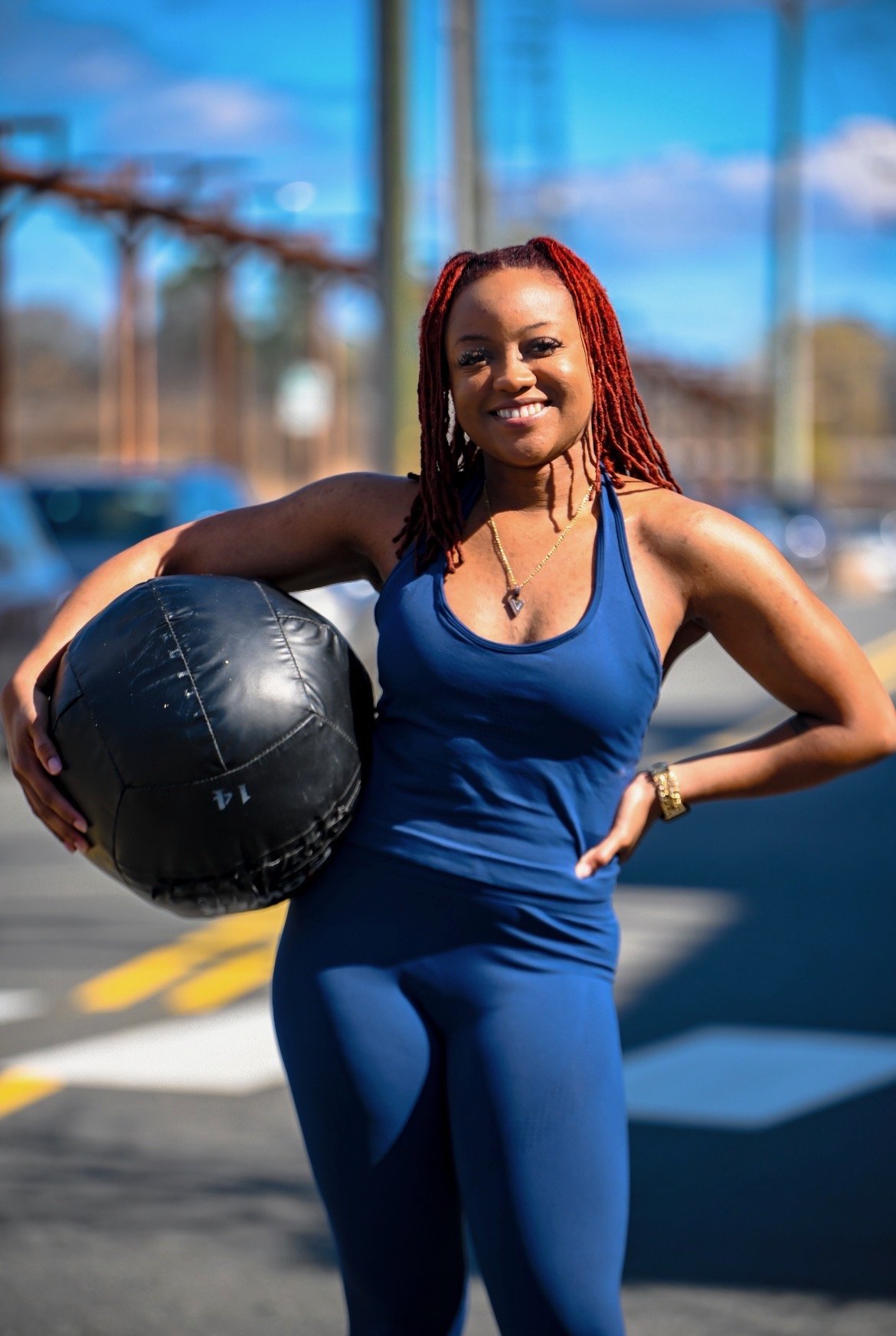 A woman wearing a blue workout outfit holding a medicine ball in a parking lot on a sunny day.