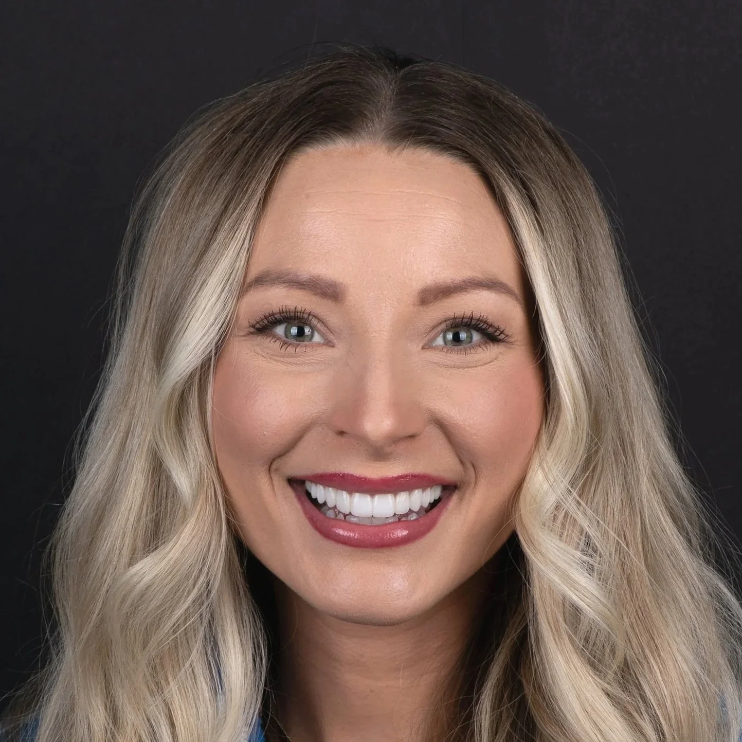 Close-up of a smiling woman with long blonde hair and blue eyes, showing teeth, against a black background.