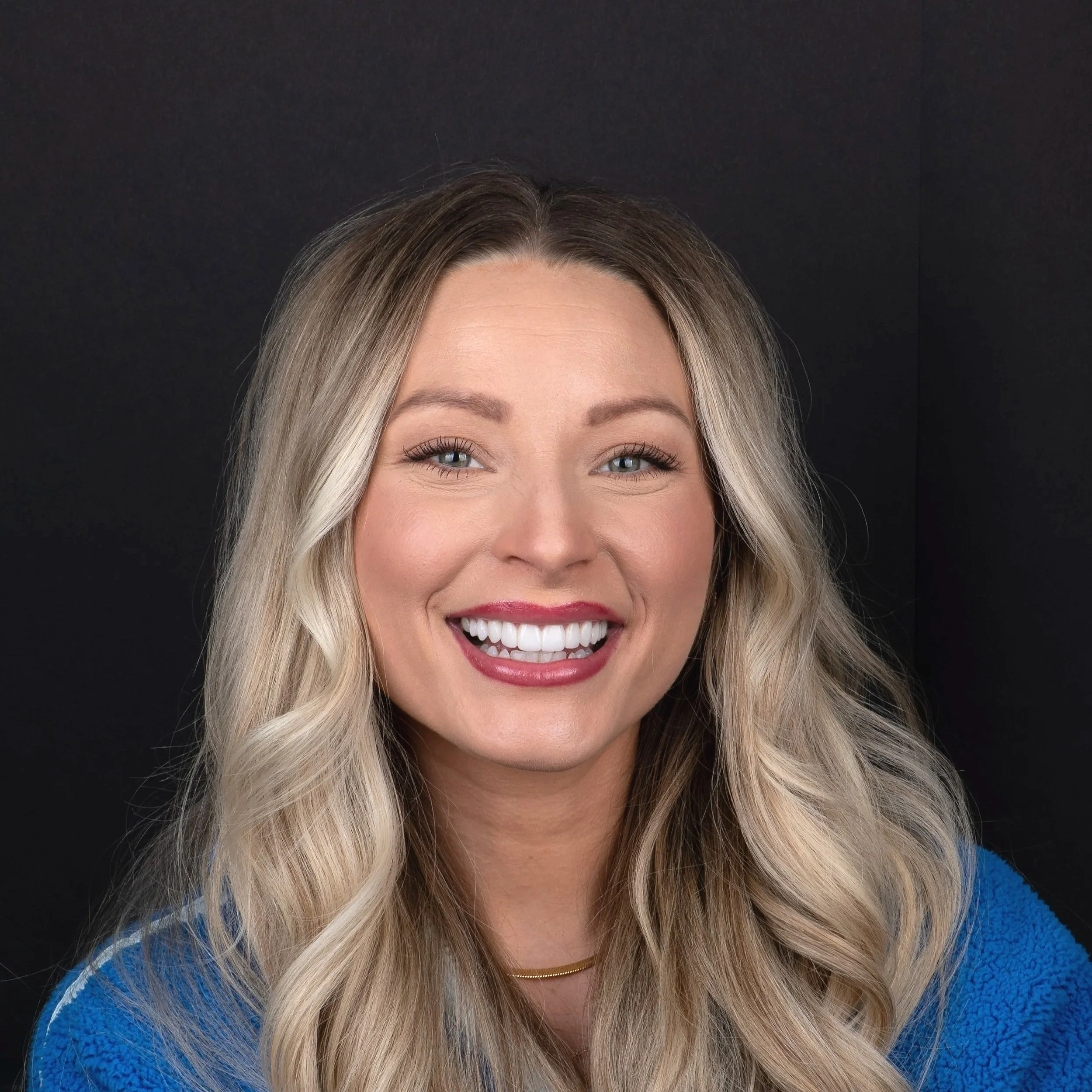 Close-up of a woman with blonde hair, blue eyes, smiling showing teeth, wearing a blue sweater, and a gold necklace against a black background.