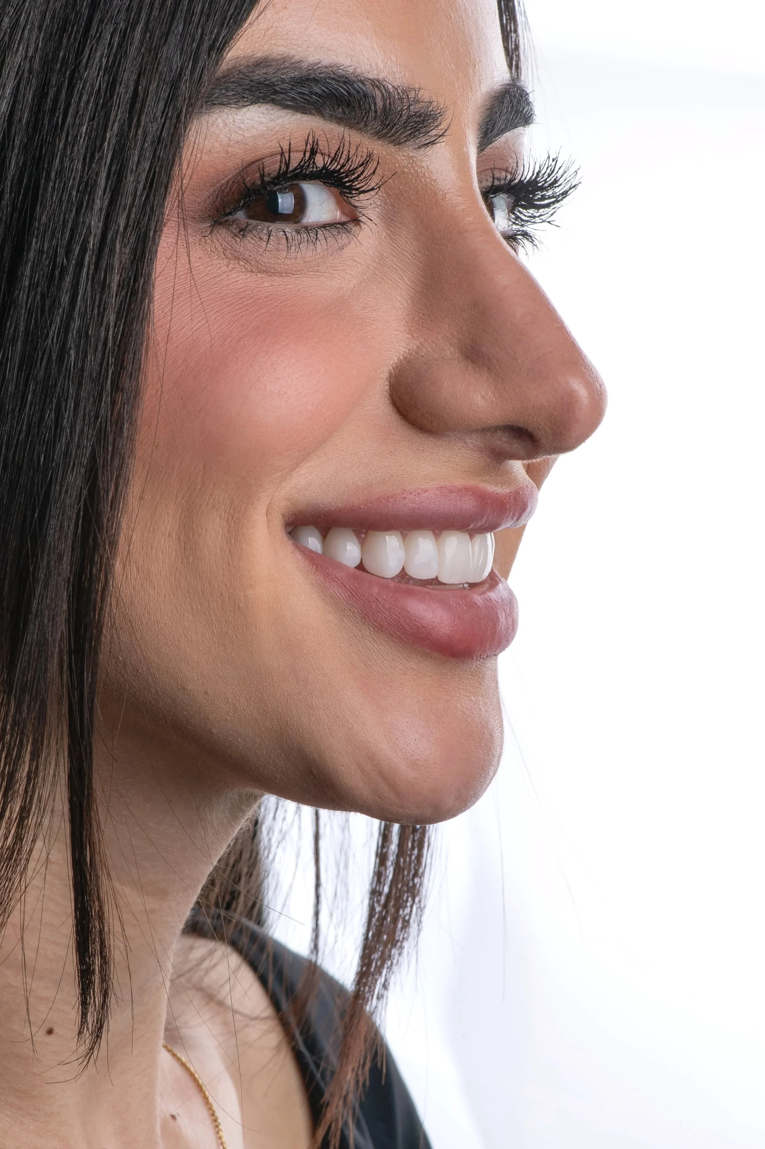 Close-up of a woman's face showing her smiling with white teeth, dark hair, and makeup, against a white background.