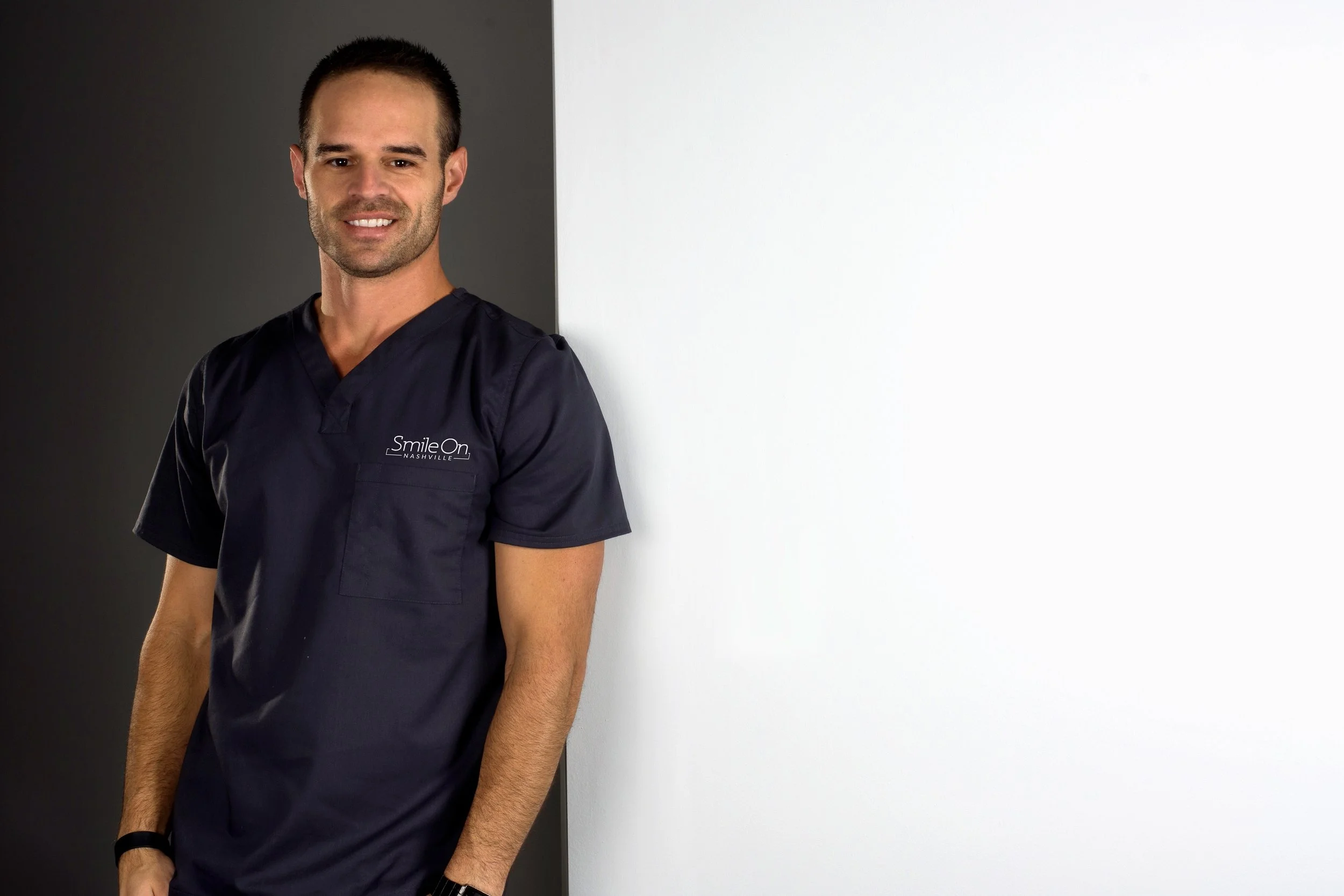 A man wearing navy blue scrubs with 'Smile On Nashville' logo on the chest, standing against a background divided into black on the left and white on the right, smiling and looking at the camera.