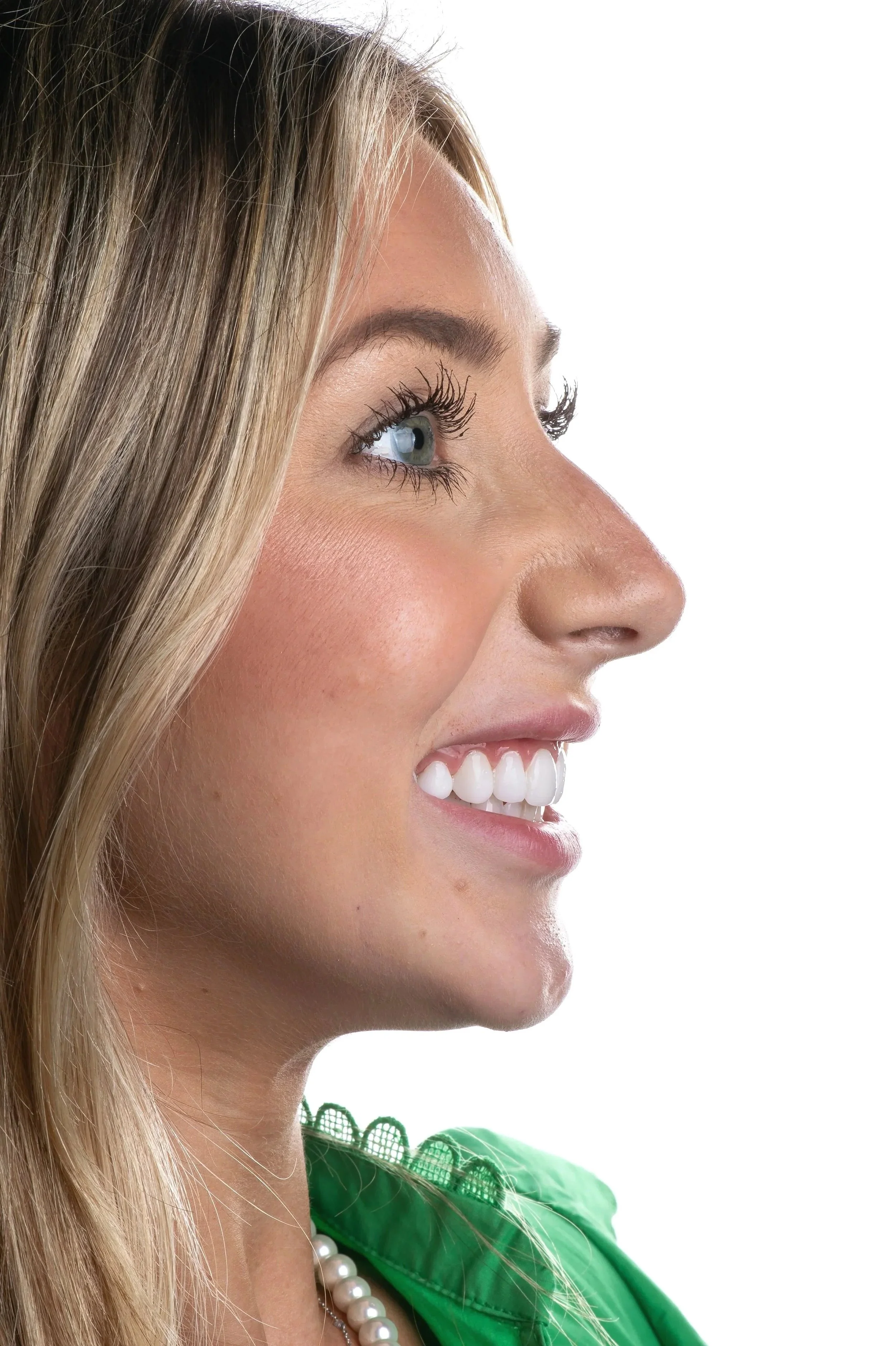Close-up of a woman's face in profile showing her eye, nose, and mouth, with blonde hair, wearing a green garment, pearl necklace, and a bright smile.
