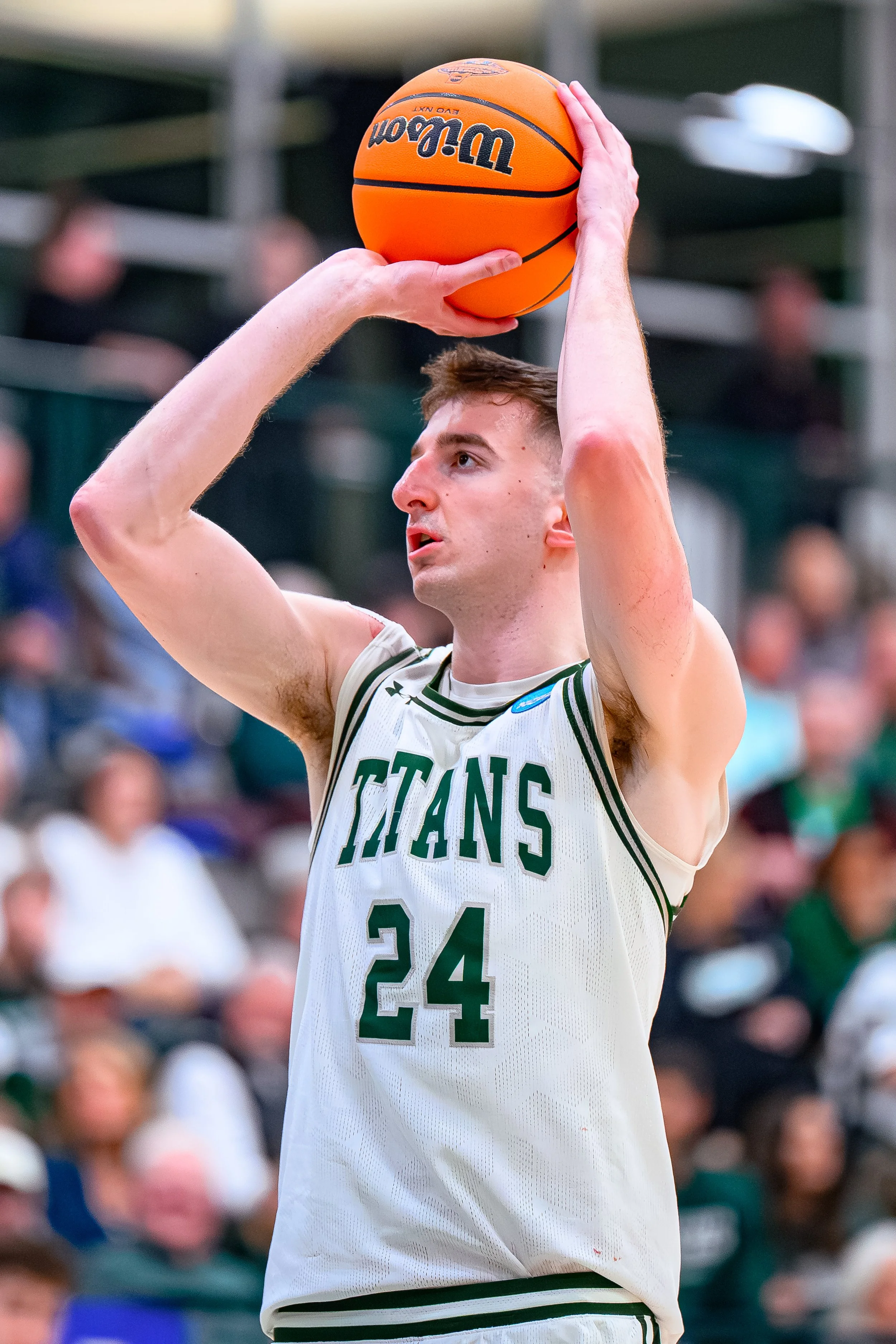 A basketball player in a white jersey with green accents and the word 'Titans' holding an orange basketball above his head, preparing to shoot during a game with spectators in the background.