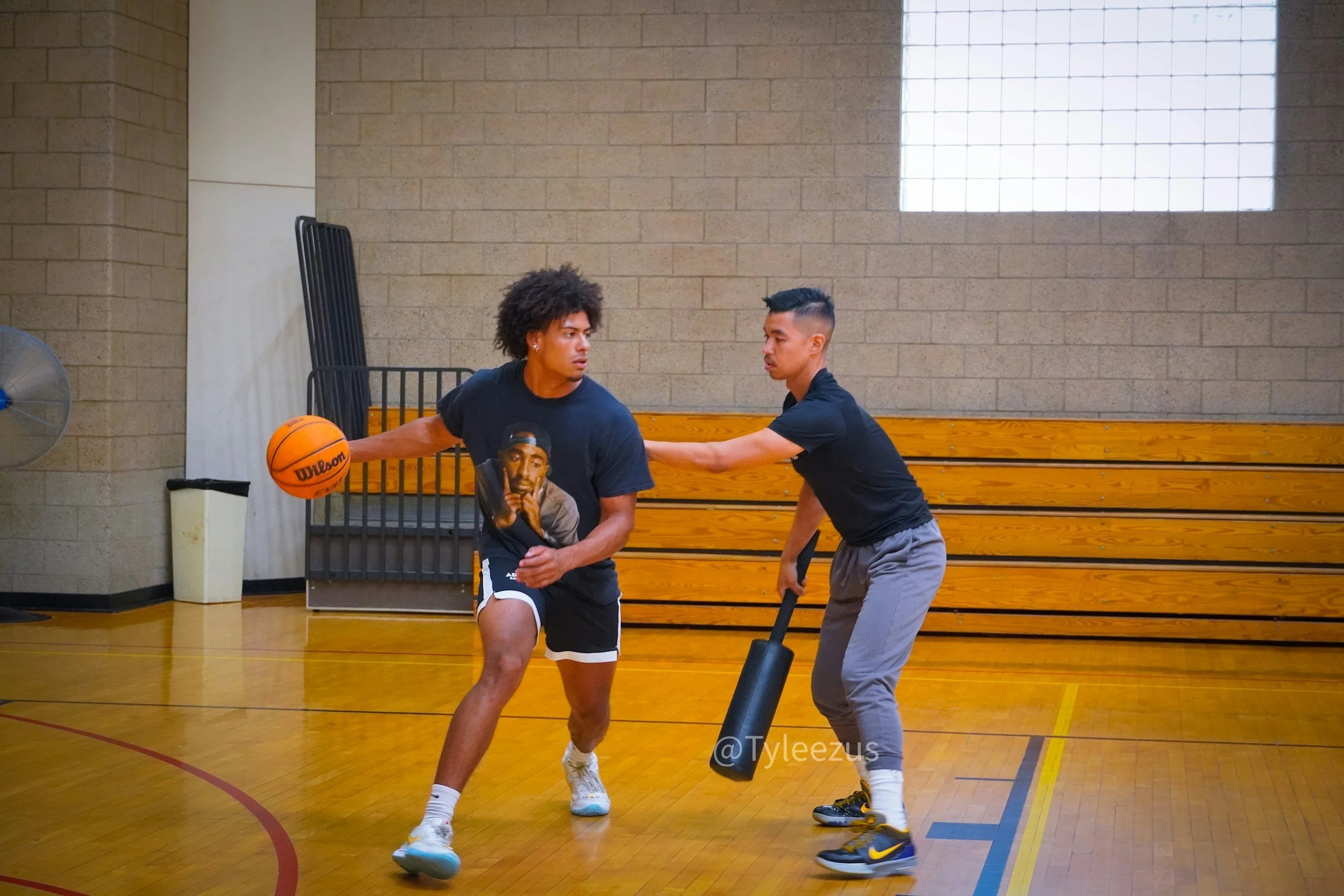 Two young men playing basketball in an indoor gymnasium, one dribbling the ball while the other defends, with wooden bleachers and a brick wall in the background.