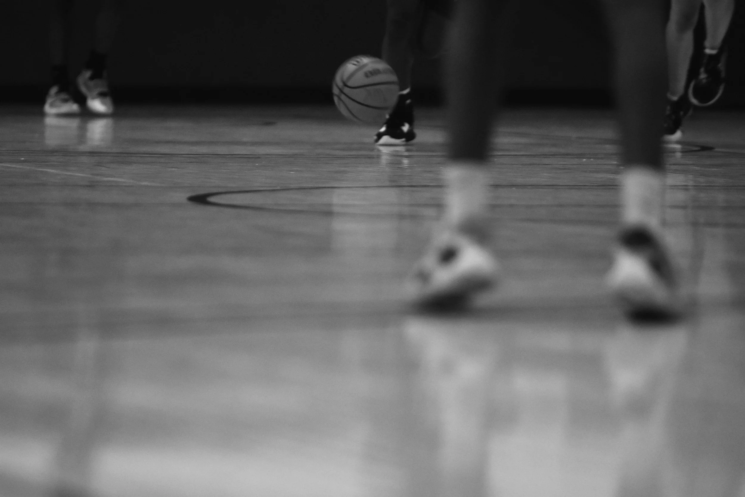 Black and white photograph of a basketball court with a basketball on the floor, amidst multiple players, some visible from the knees down.