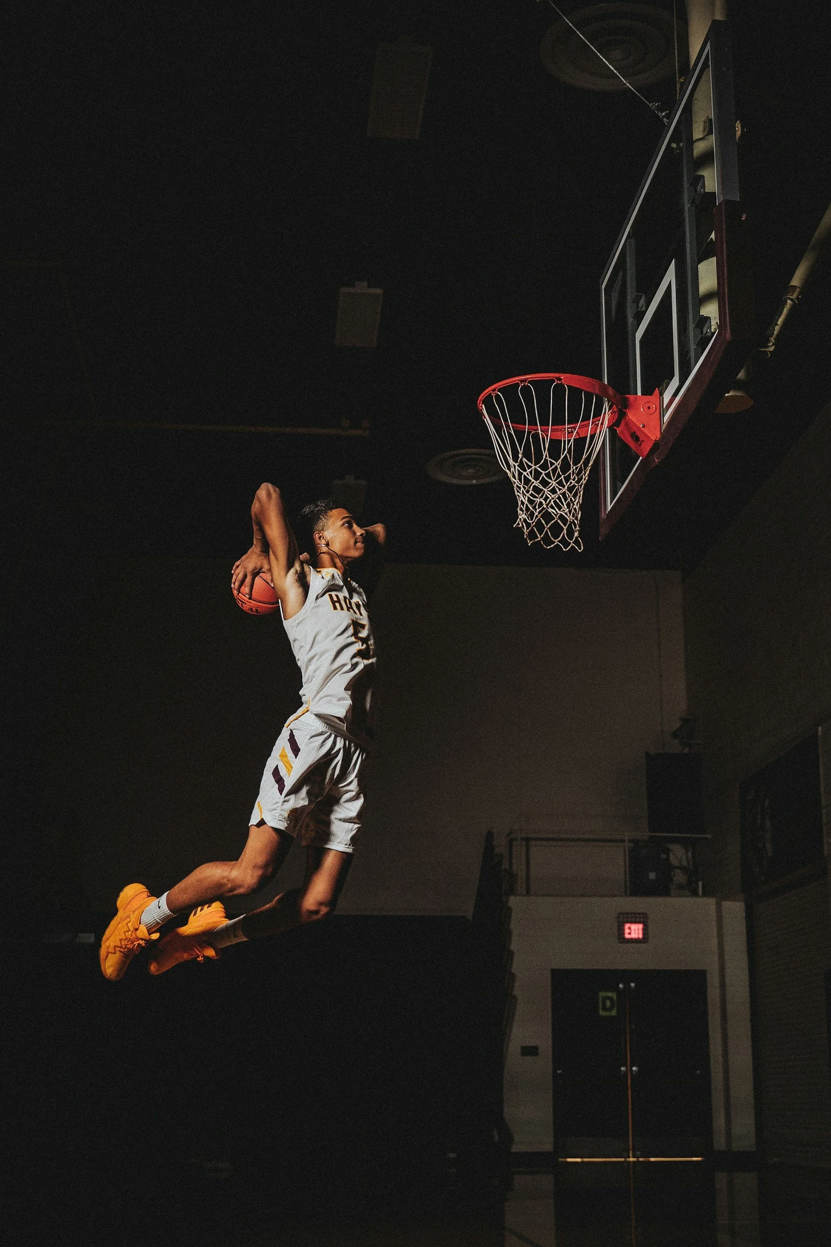 A basketball player is jumping towards the hoop for a dunk shot in an indoor court, with dark lighting and a black ceiling.