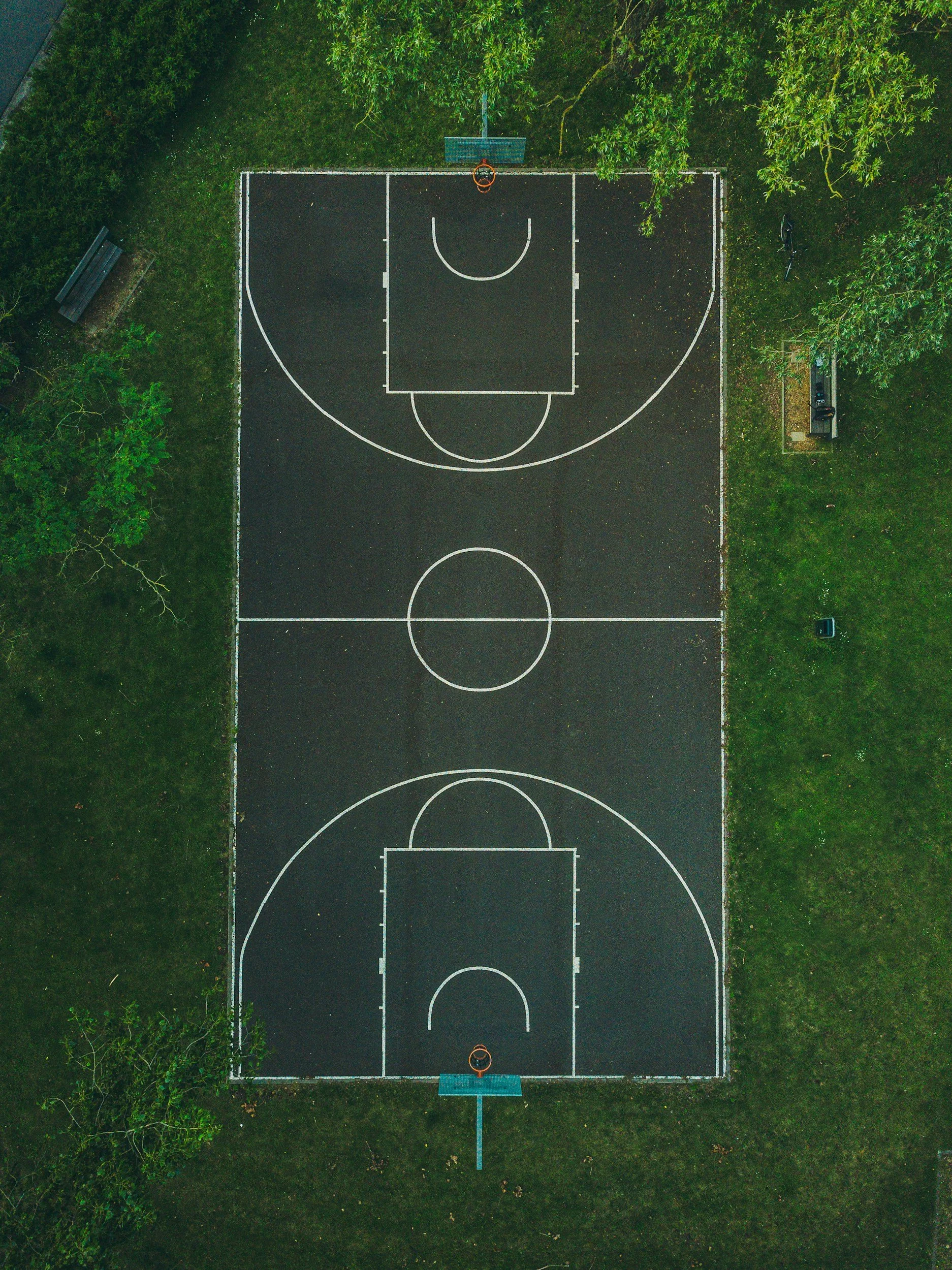 An aerial view of a basketball court situated outdoors, surrounded by green grass and trees. The court has painted white lines, a basketball hoop at each end, and a basketball net visible at the top of the image.
