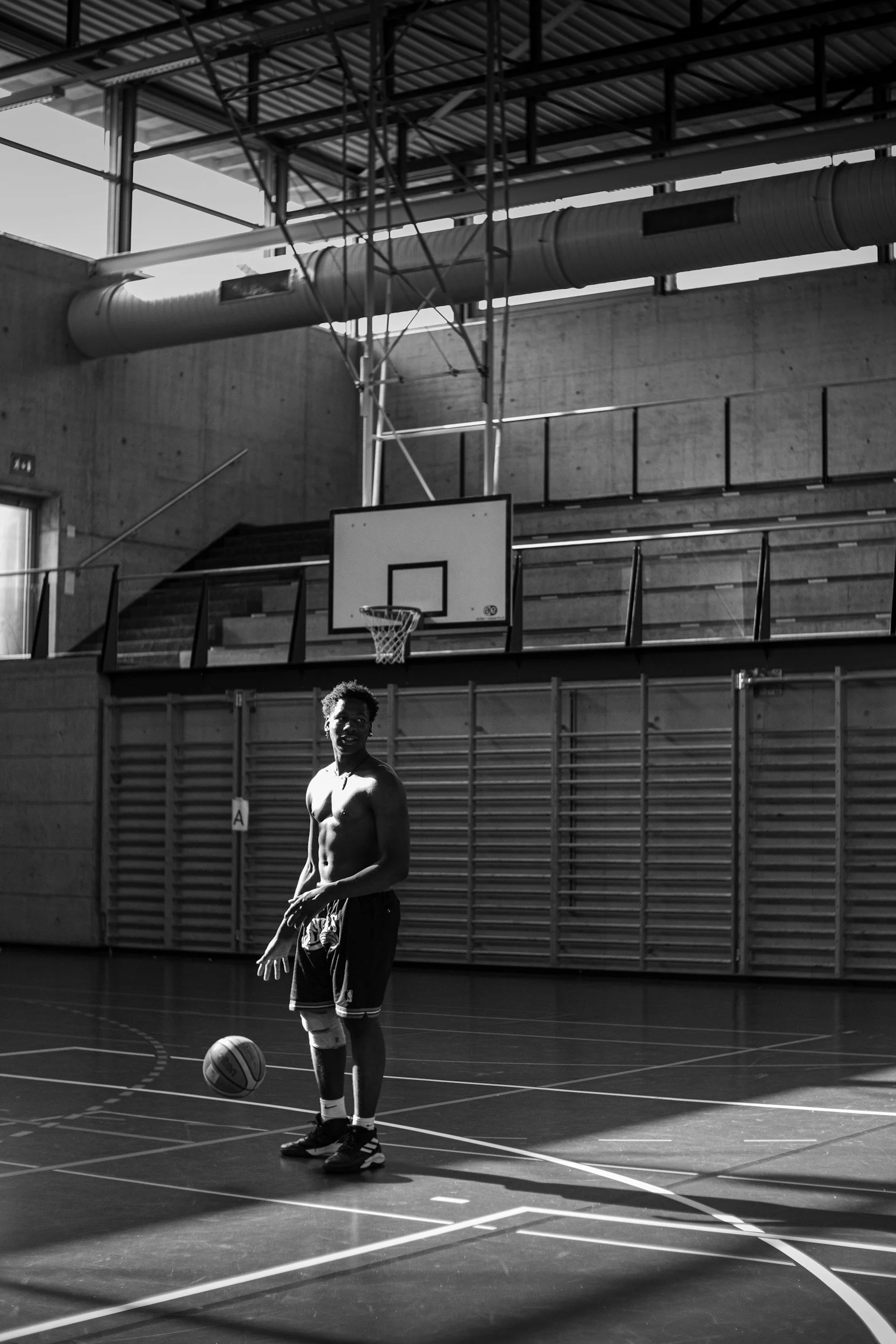A young man standing on an indoor basketball court, holding a basketball with a ball at his feet. The gym has high ceilings, a basketball hoop, and a large ventilation pipe above. The image is in black and white.