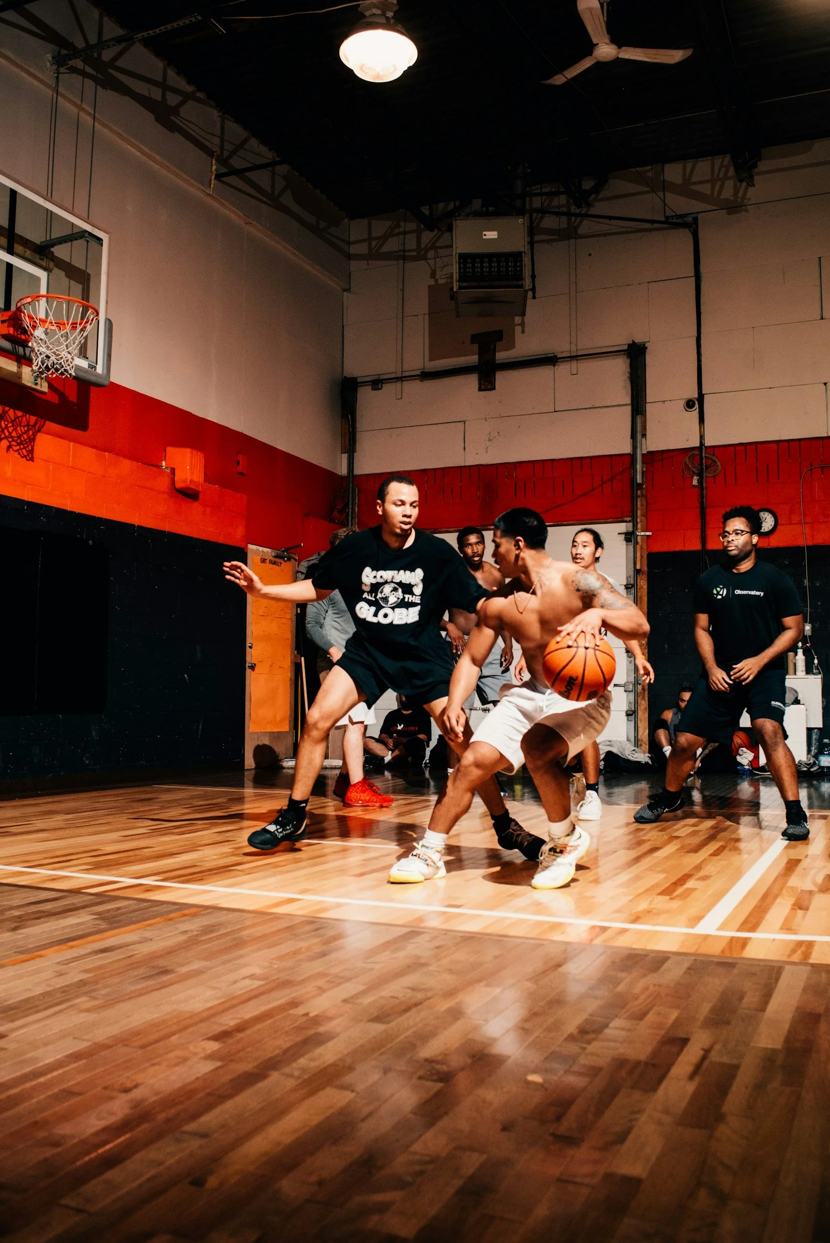 Young men playing basketball in an indoor gym, with one player dribbling the ball while others defend and watch.