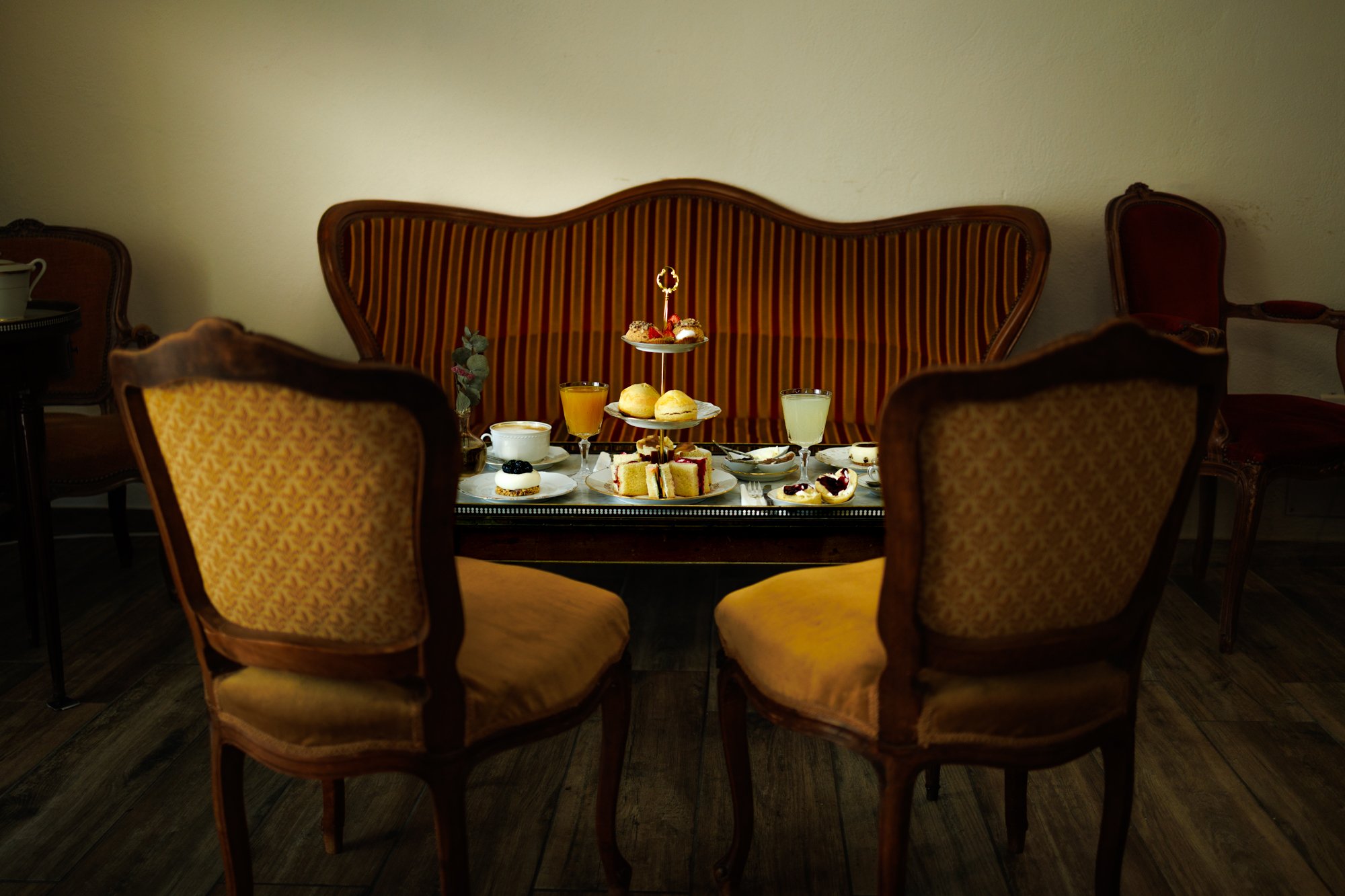 Salon de thé cosy à Montpellier,  "Chez Apolline", proposant un Tea Time à l'anglaise. La photo montre une table bouillote dressée avec des fauteuils et une banquette d'époque, un présentoir à trois niveaux rempli de pâtisseries.