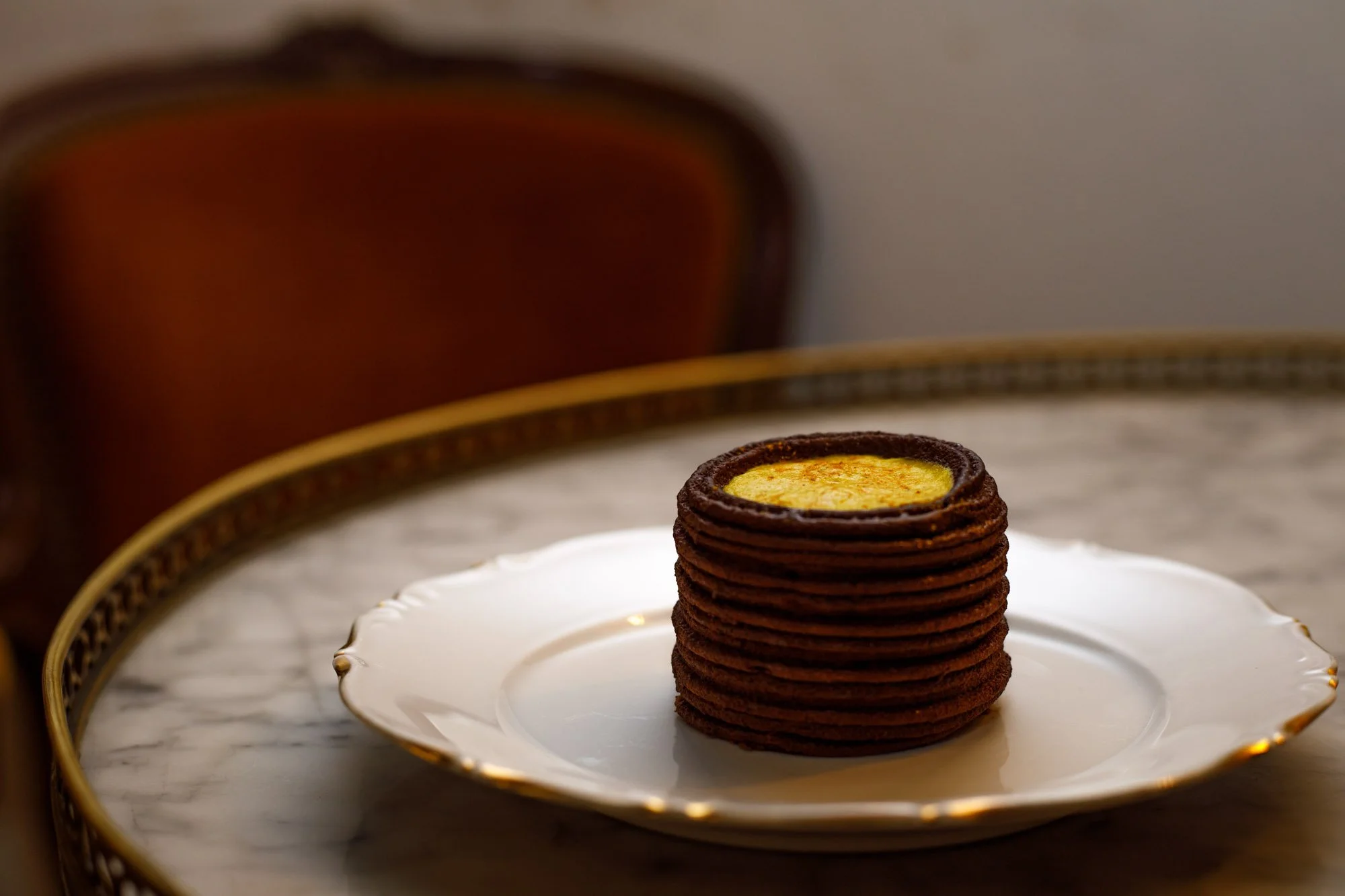 Entremet au chocolat et coulis jaune orangé dressé sur une assiette en porcelaine posée sur une table ronde en marbre