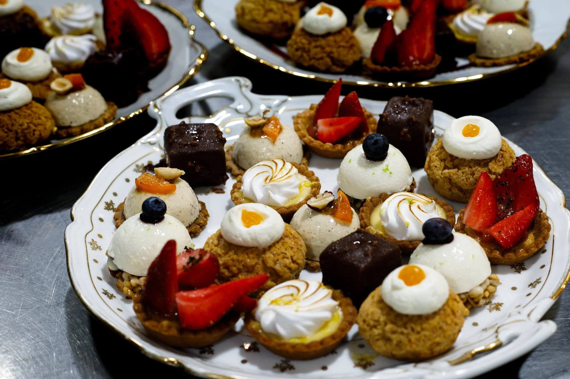 Assortiment de mini tartes et desserts sur une assiette en porcelaine ornée de motifs dorés, incluant des tartes aux fruits, des bouchées chocolatées, des mignardises avec de la crème et des fruits.