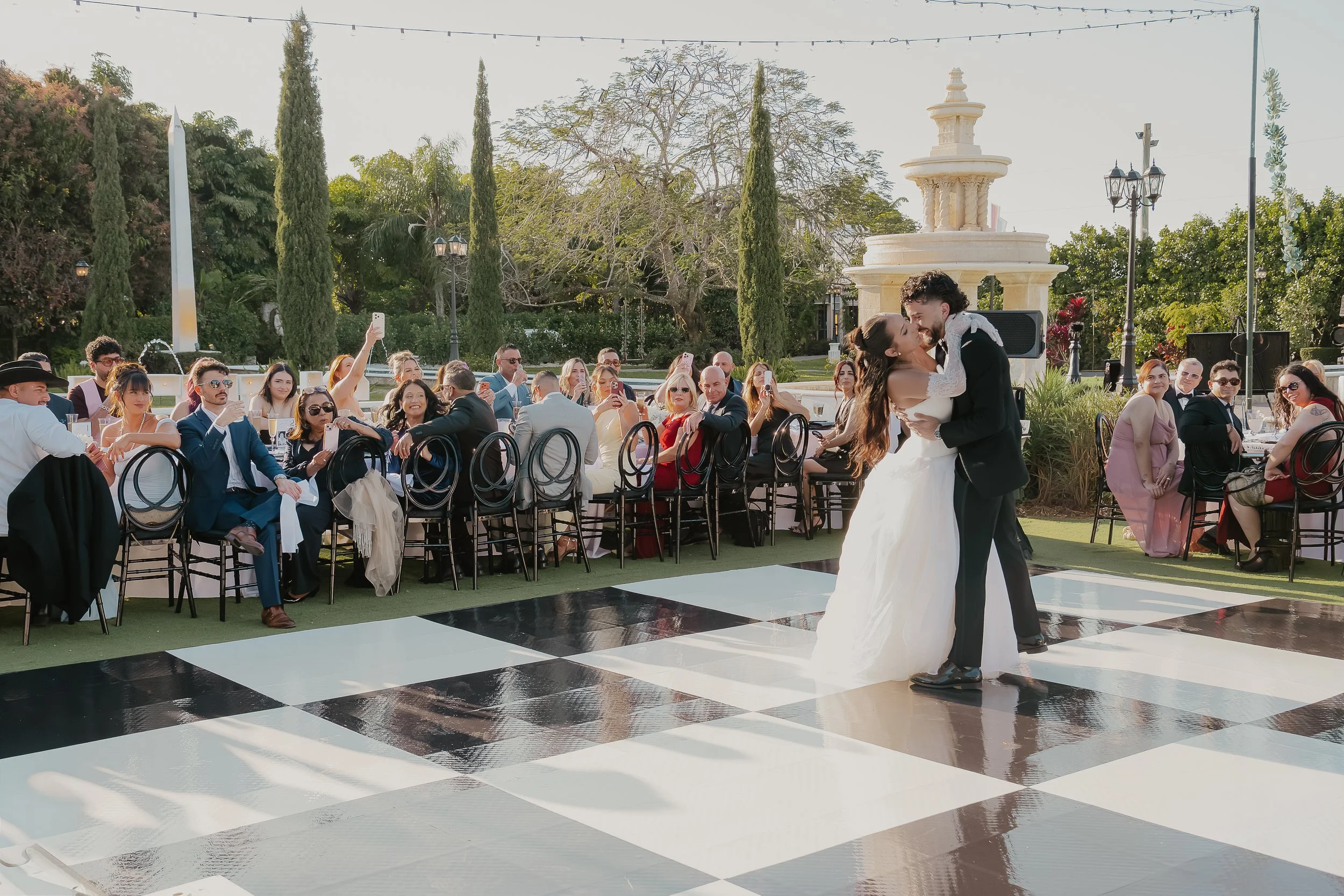 A couple is dancing in front of guests seated outdoors at a wedding reception. The guests are taking photos and watching, with a backdrop of trees, a fountain, and string lights.