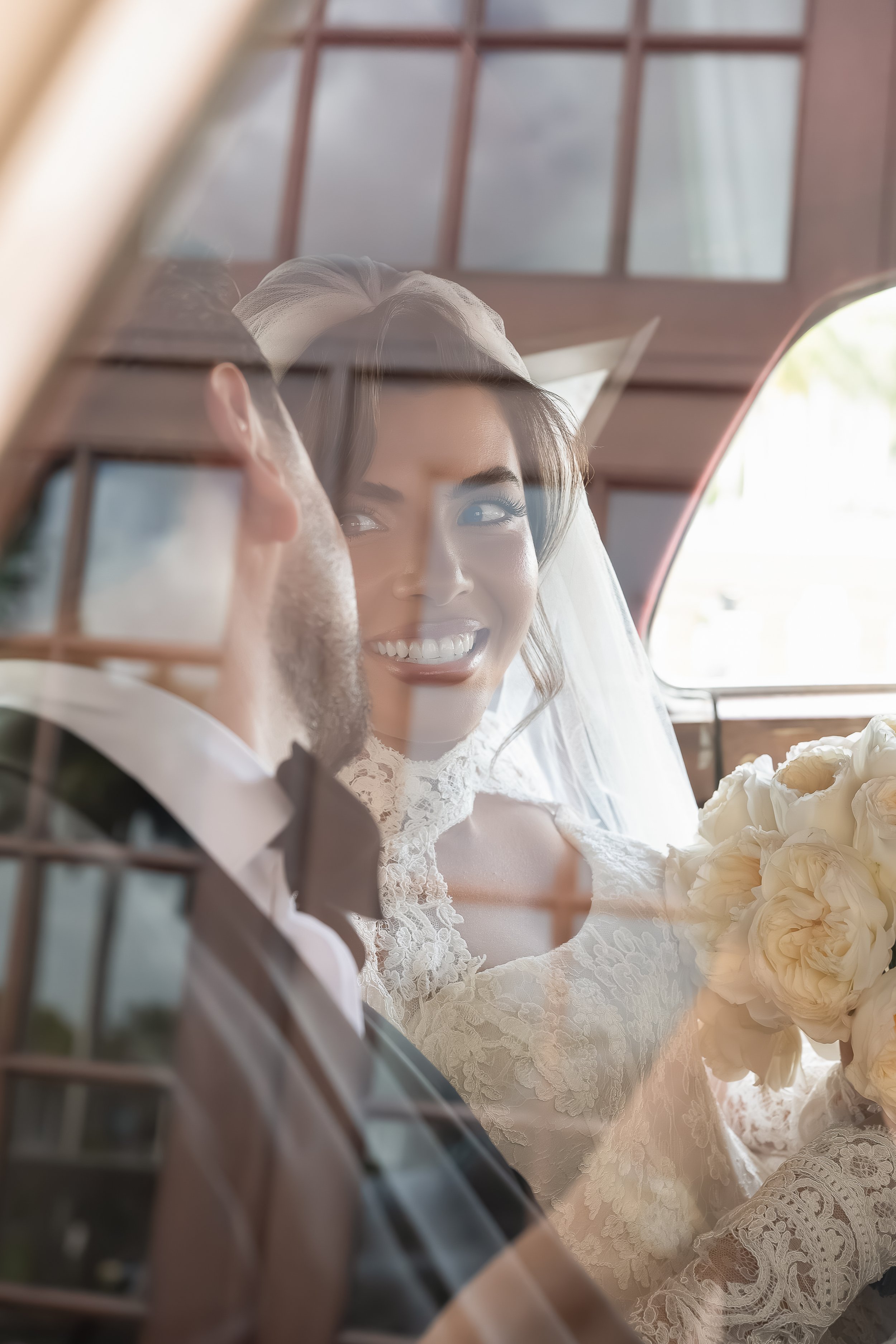 A smiling bride in a lace wedding dress holding a bouquet of white flowers, seated next to a groom in a suit, as seen through the reflection of a glass window with grid pattern in a wooden setting.