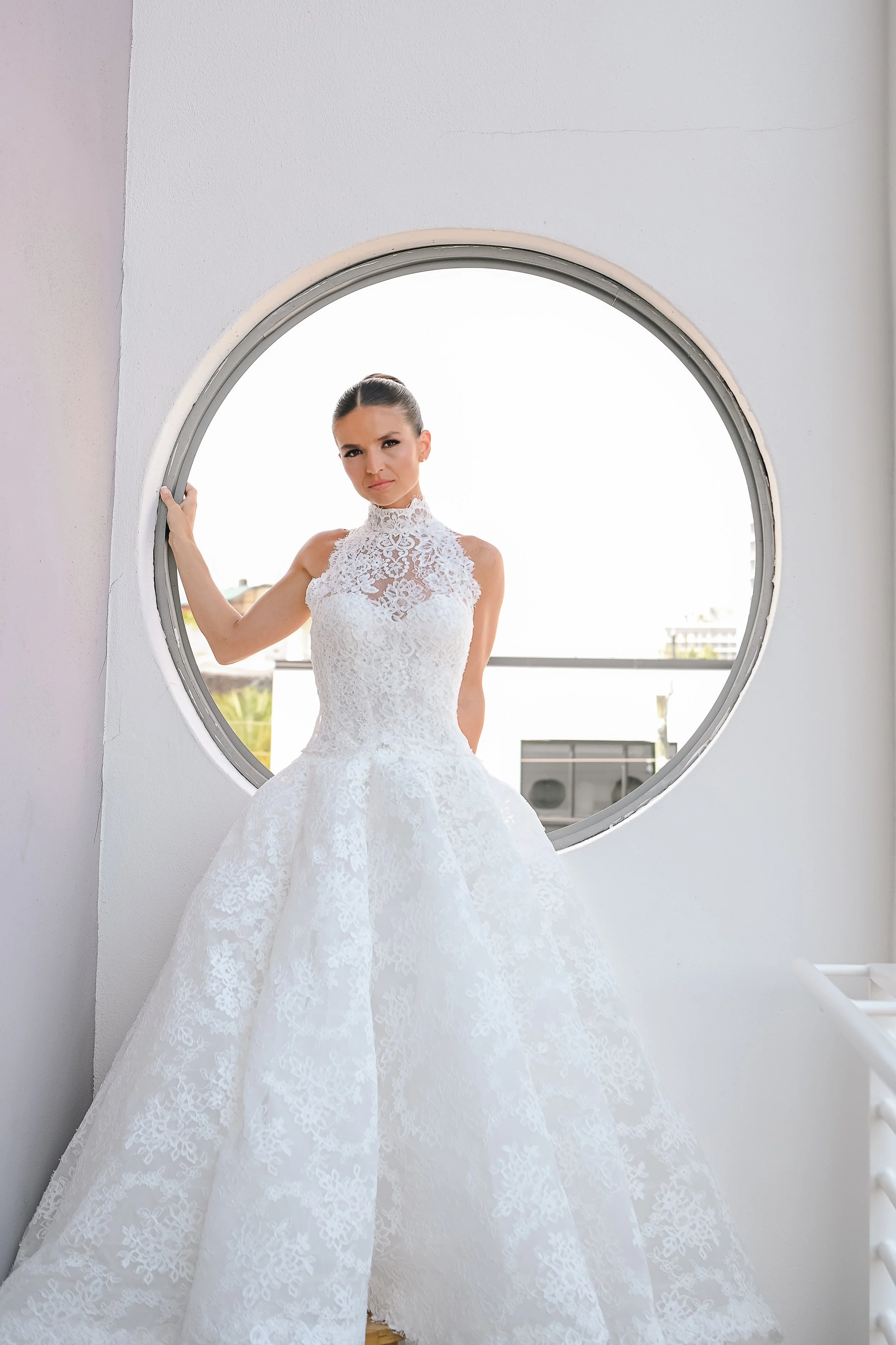 A woman in a white lace wedding dress standing in front of a circular window, holding the window frame with one hand, with a cityscape visible in the background.