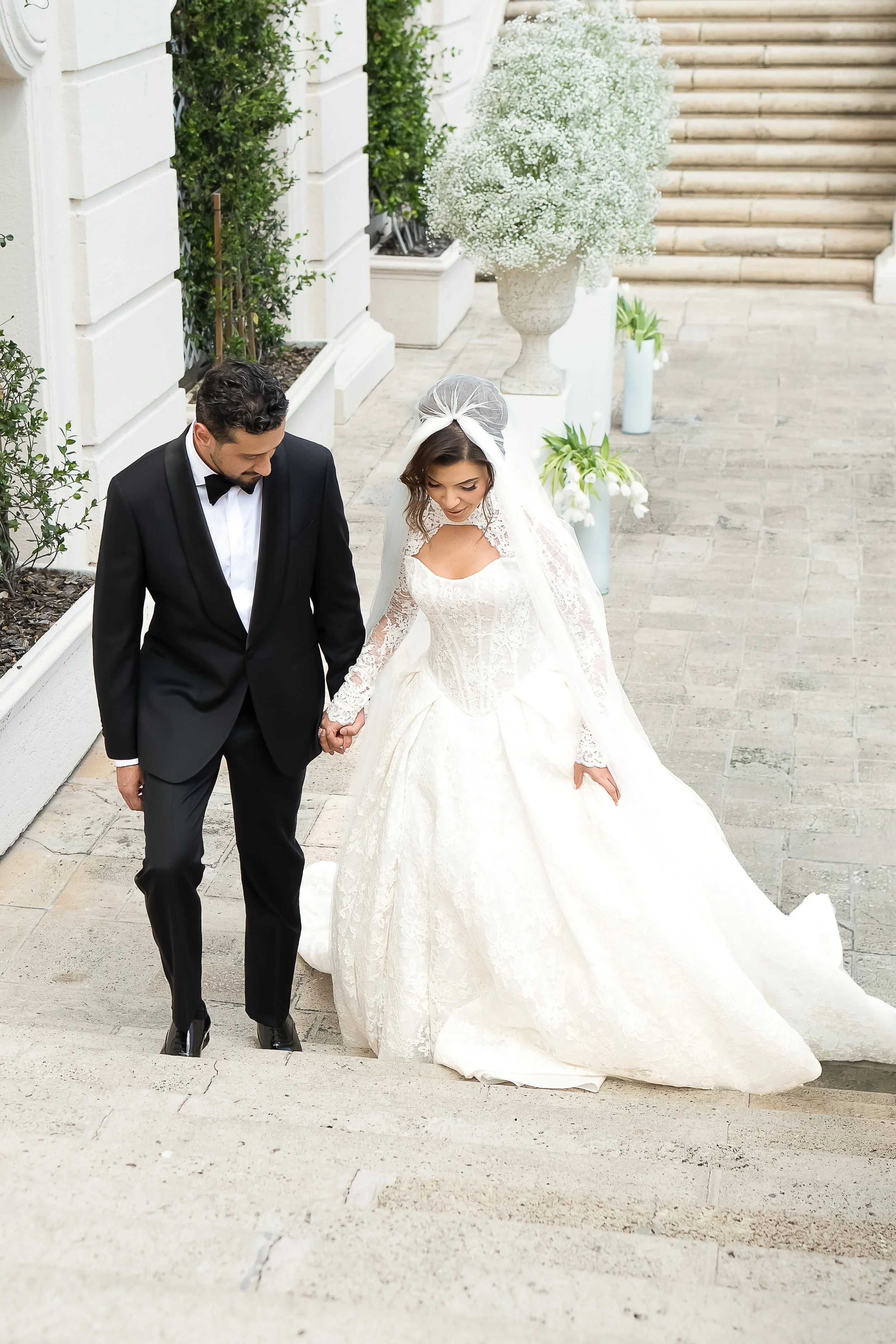 A bride and groom walking down stairs, holding hands, with the bride wearing a white wedding gown and veil, and the groom in a black tuxedo, surrounded by decorative plants and staircase.