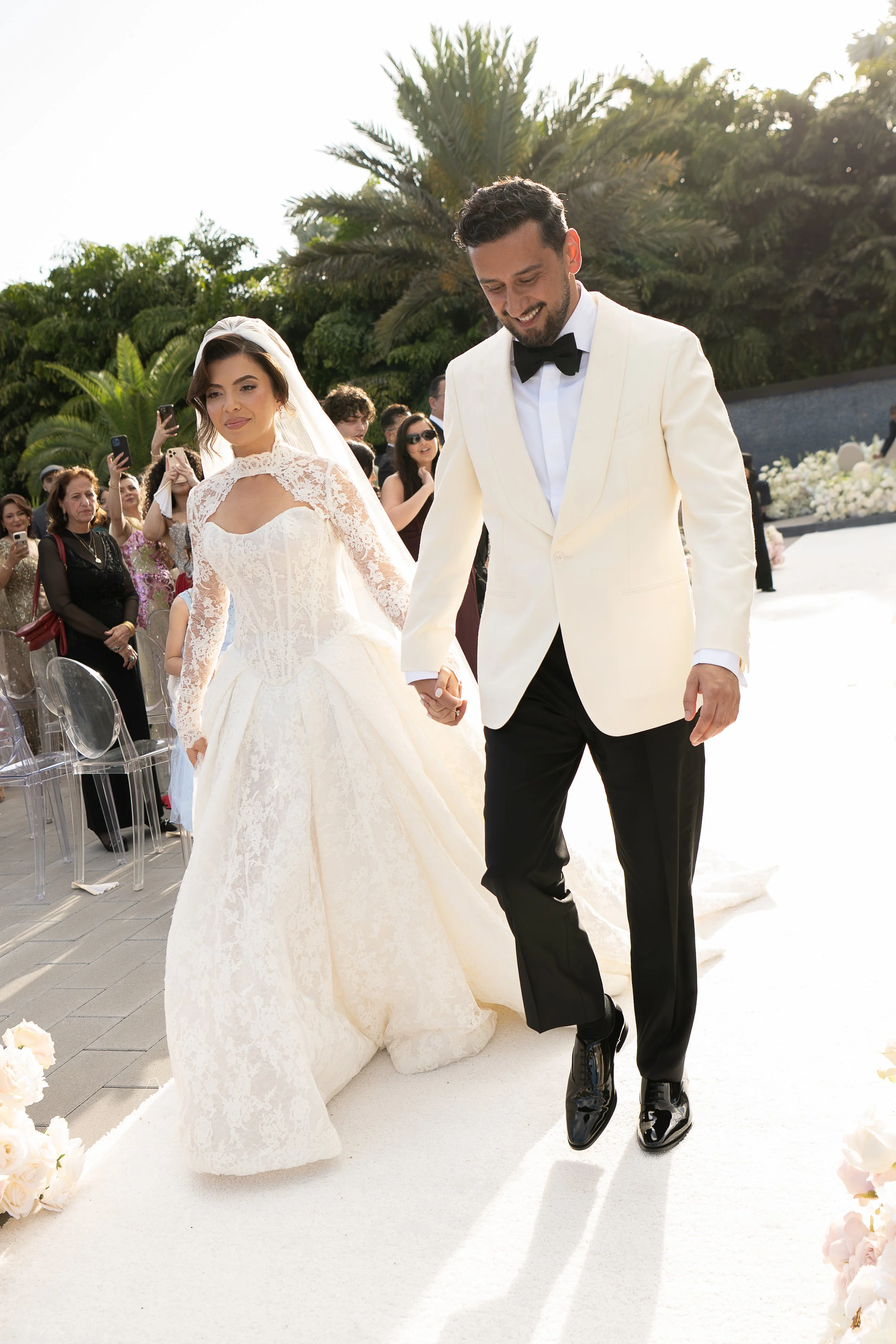 A bride and groom holding hands during their wedding ceremony outdoors, with guests watching in the background and greenery behind them.
