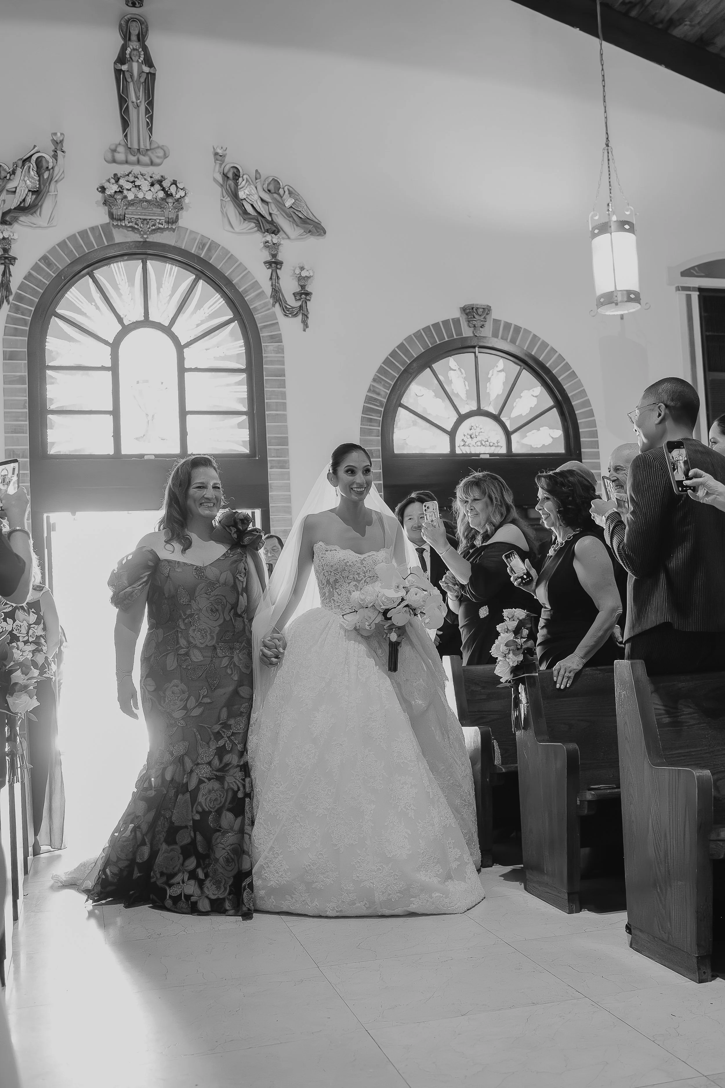 A bride in a wedding gown holding a bouquet, standing with a woman in a floral dress inside a church, with a group of people taking photos. The church has arched windows with stained glass and religious statues and decorations on the wall.