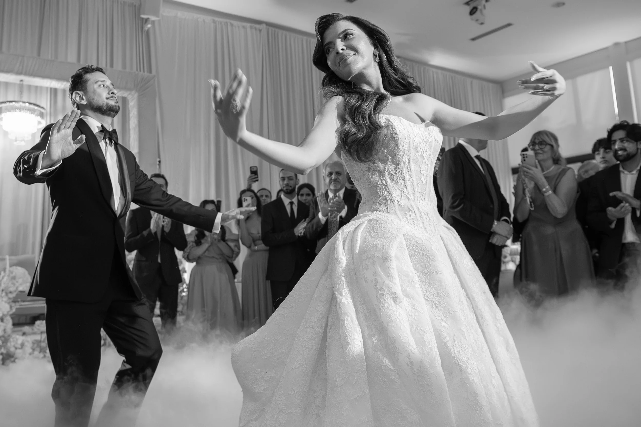 A bride and groom dancing at their wedding reception with guests watching and taking photos. The bride is wearing a white wedding gown and the groom is in a black tuxedo.