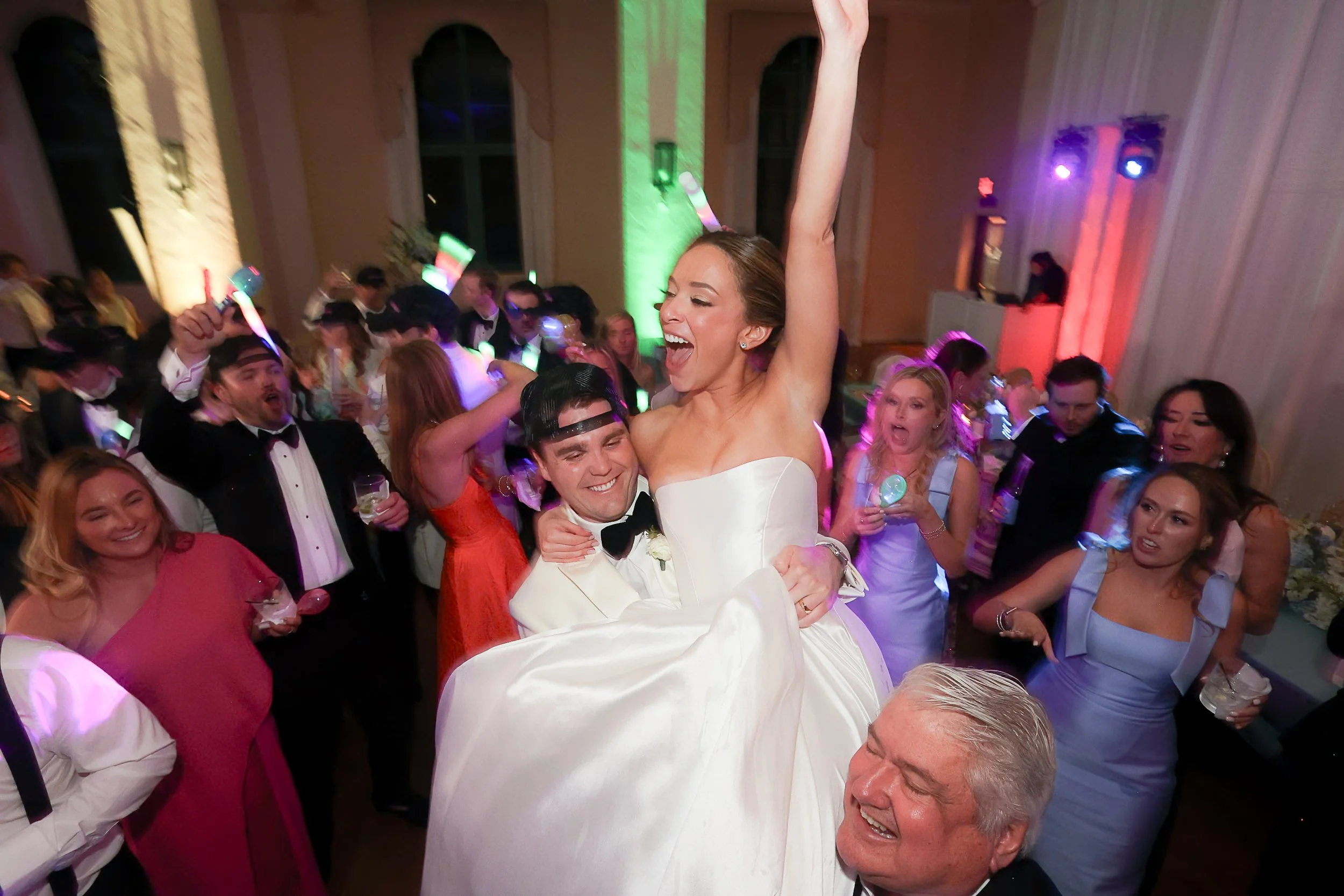 A woman in a white wedding dress raises her arm in celebration, joyfully smiling while being lifted by a man in a tuxedo. They are surrounded by guests dancing at a lively wedding reception with colorful lighting and excited expressions.