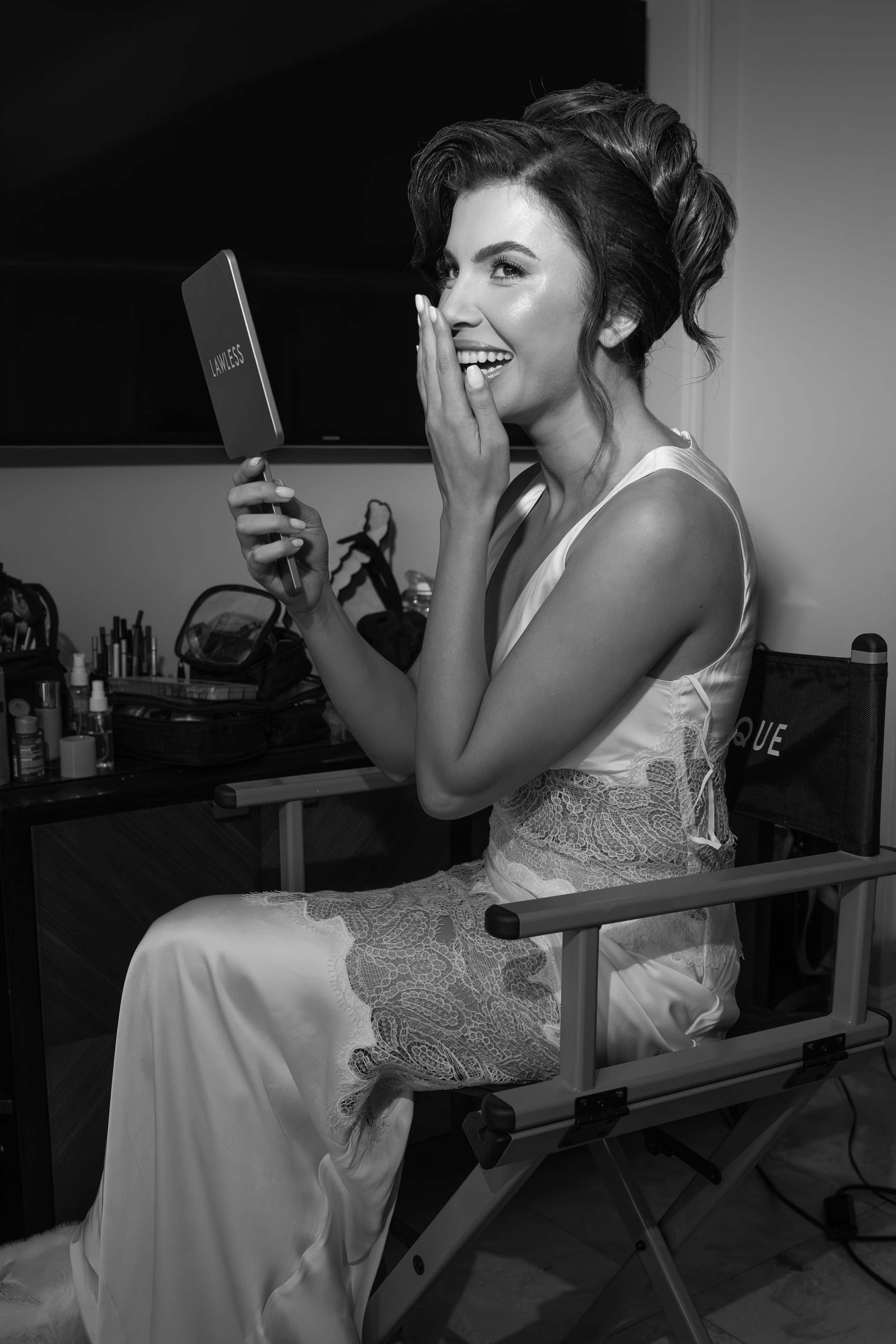 A woman in a satin and lace dress sitting in a makeup chair, looking at a handheld mirror with the word 'LAWLESS' on the back, smiling and covering her mouth with her hand, surrounded by makeup products.
