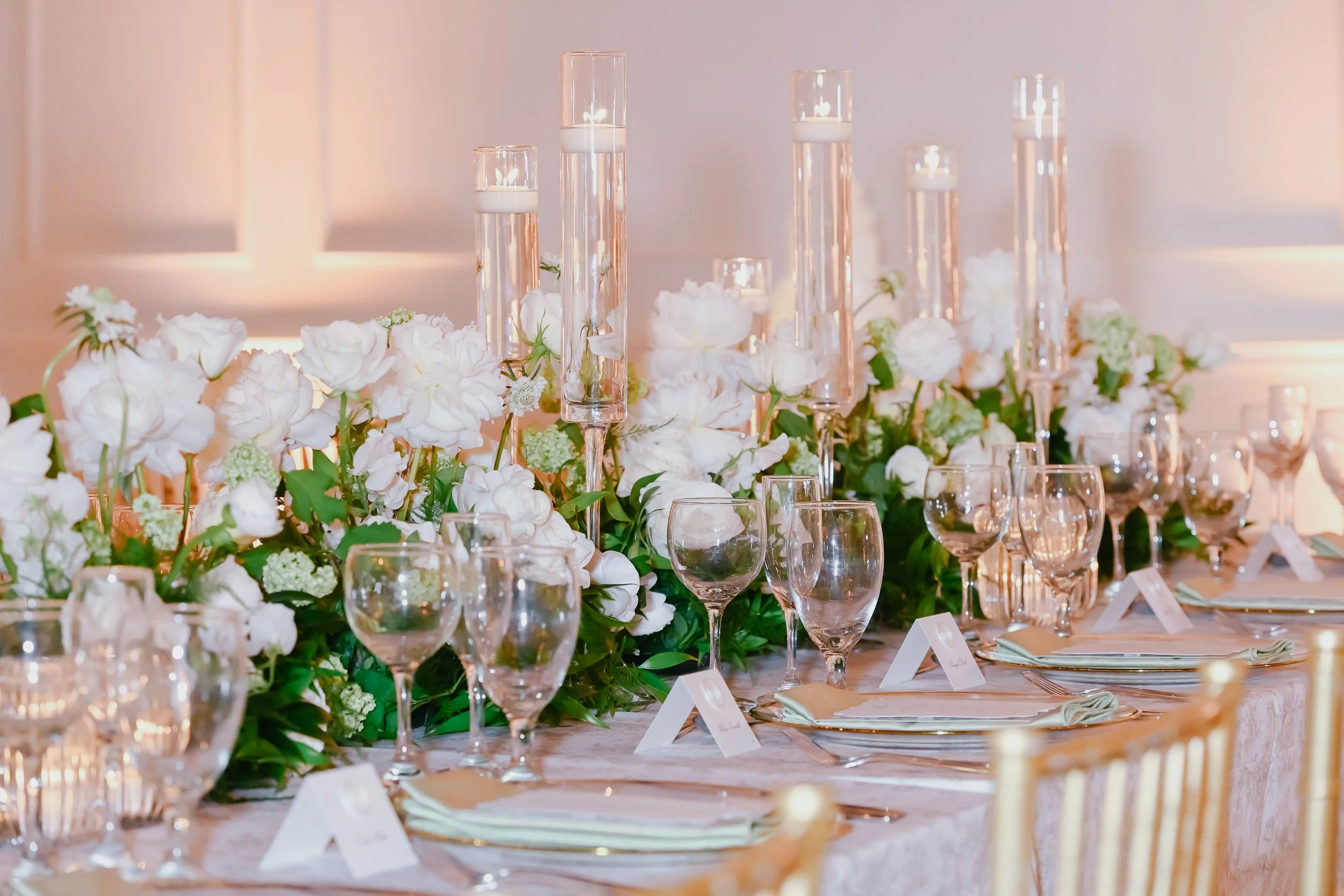 Elegant banquet table decorated with white flowers and tall glass candle holders with white candles, glassware, and place cards.