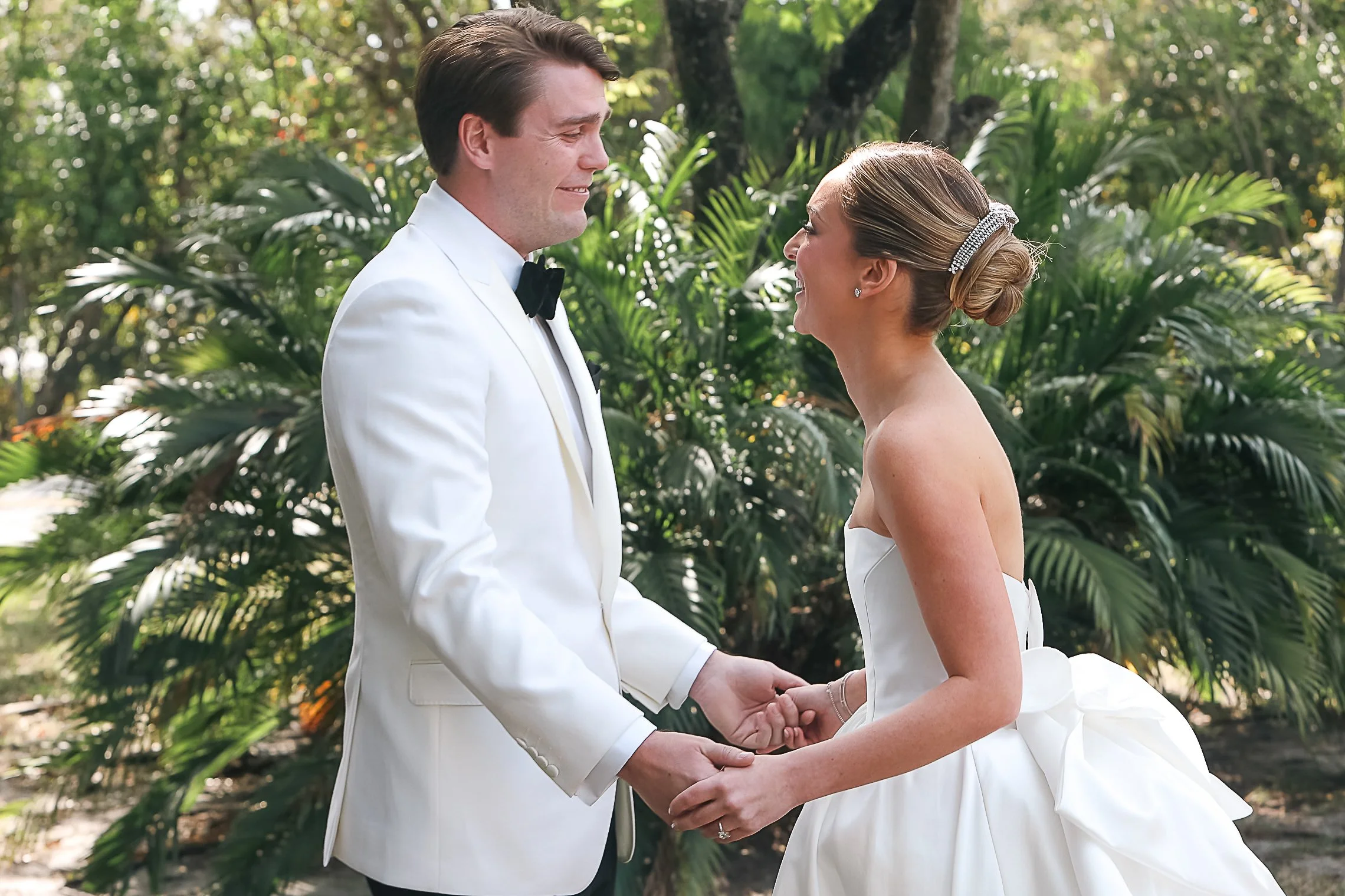 A bride and groom holding hands and facing each other outdoors in a lush green setting during their wedding ceremony.