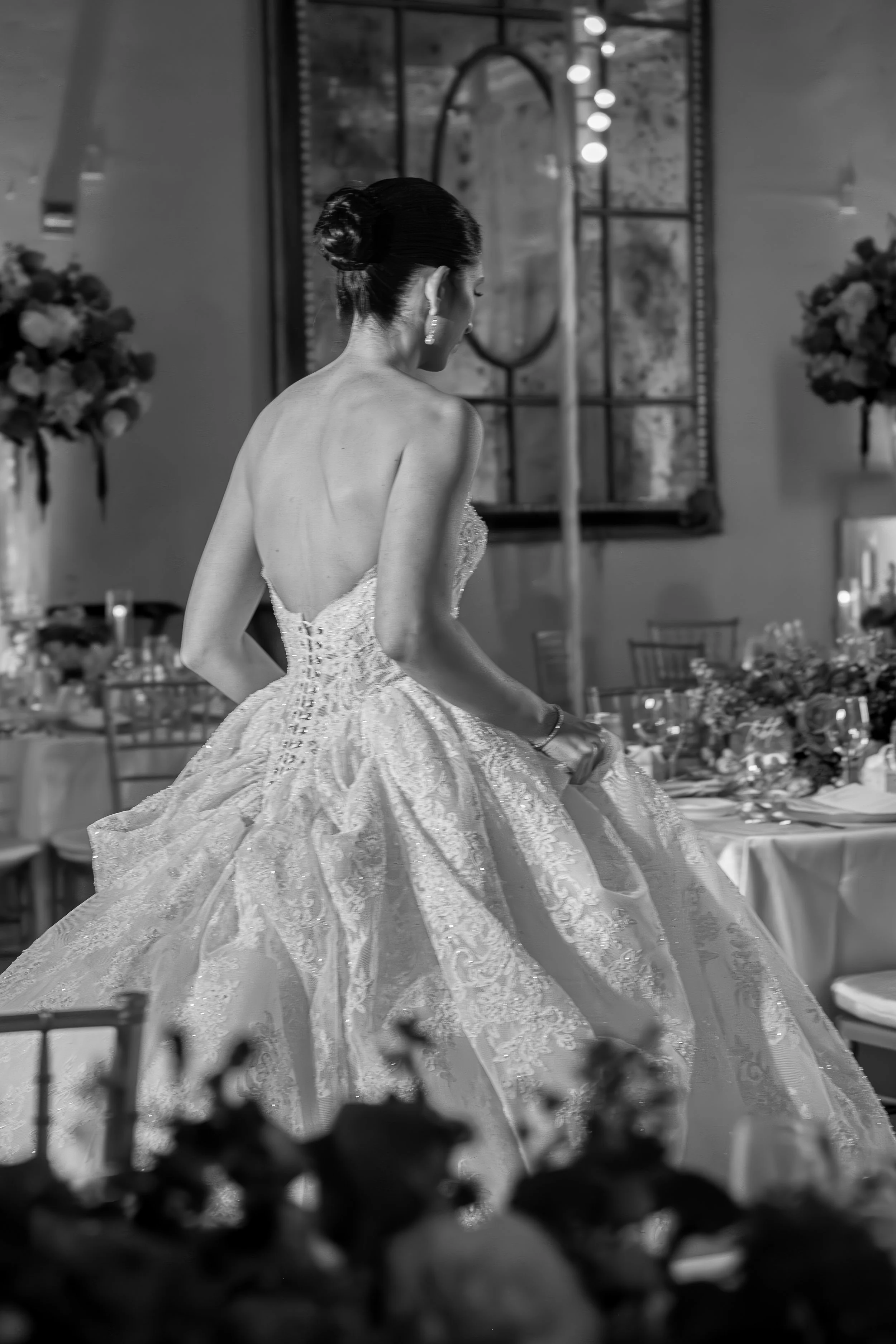A woman with dark hair styled in an elegant updo, wearing a strapless, ornate wedding gown with intricate lace detailing, captured from behind in a wedding reception setting with floral arrangements and tables visible in the background.