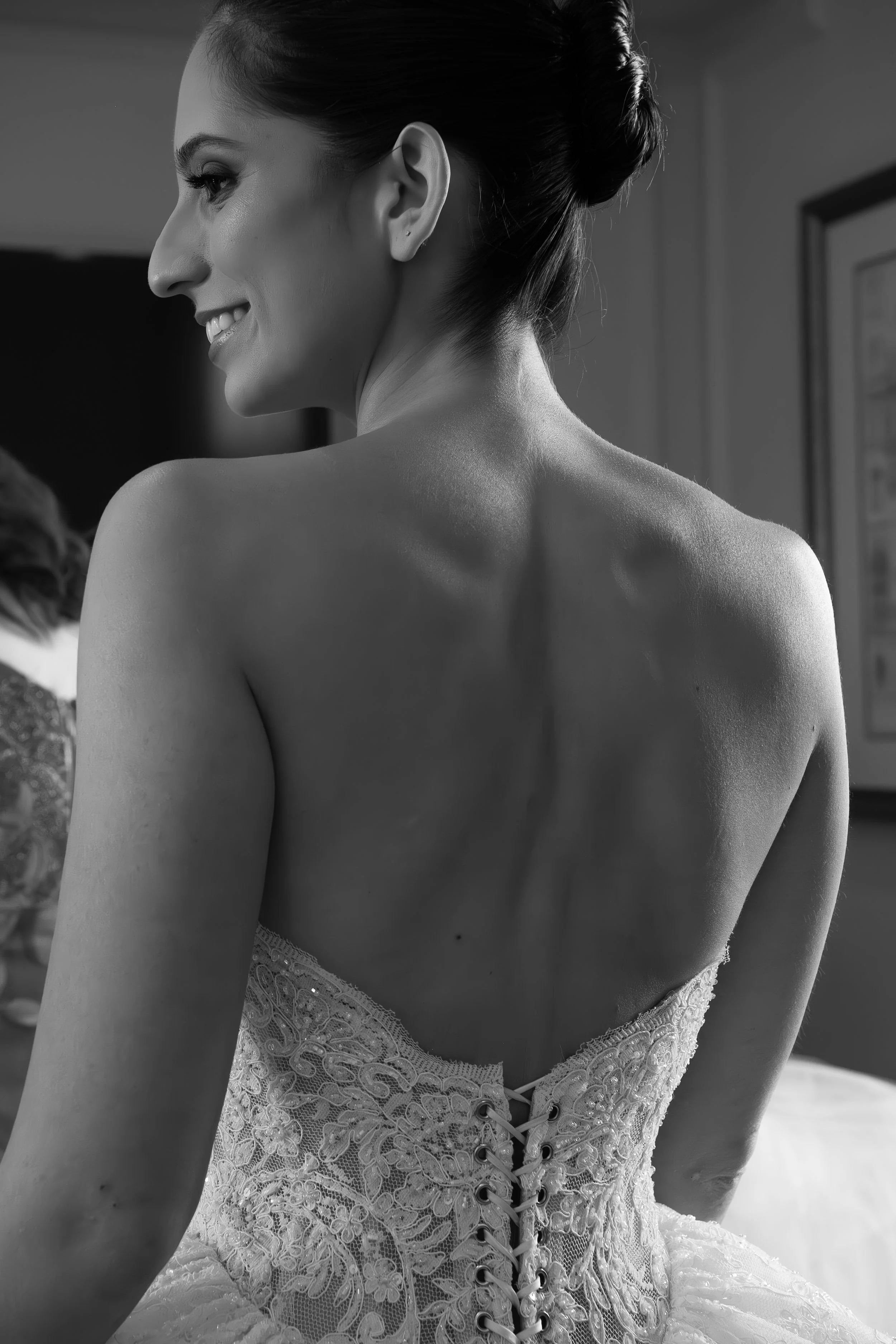 A woman with dark hair styled in an elegant bun, smiling and wearing a lace wedding dress with intricate embroidery and lacing at the back, captured in black and white.