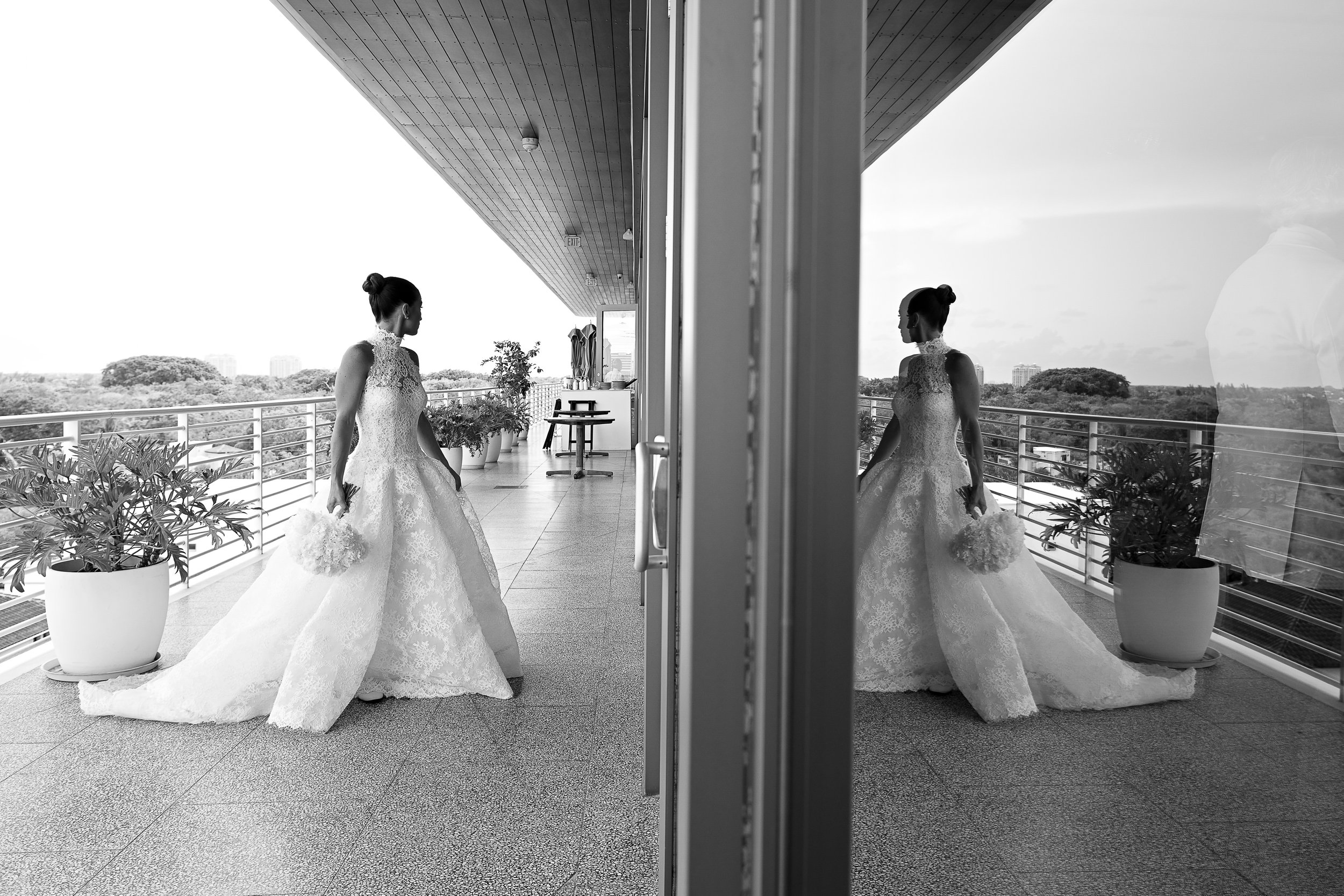 A mirror reflecting two women in wedding dresses holding flowers on a balcony with potted plants and city views.