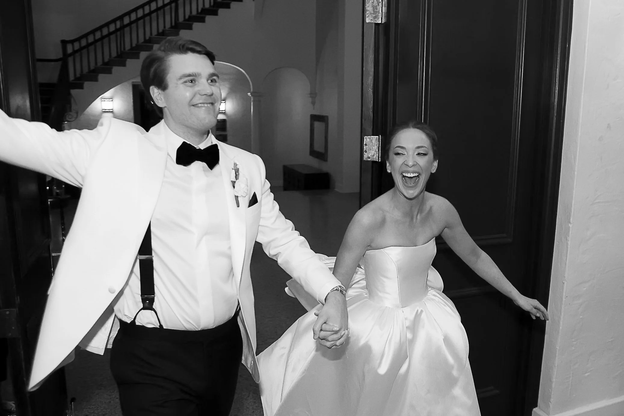 A joyful bride and groom holding hands, celebrating their wedding in an indoor setting, with the groom dressed in a tuxedo and the bride in a strapless wedding gown.