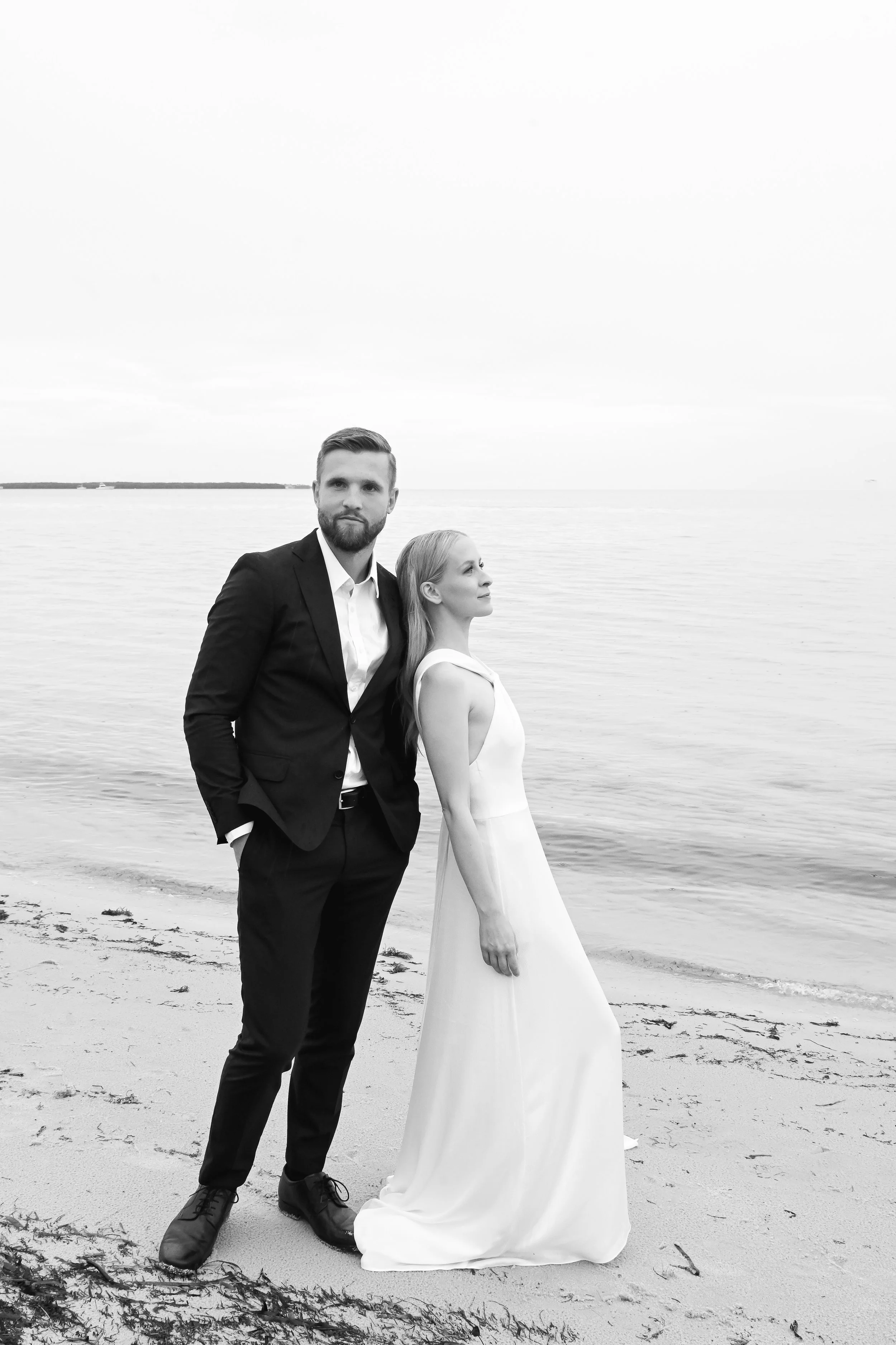 A black-and-white photo of a man and woman standing on a beach by water. The man is in a dark suit with a white shirt, hands in pockets, and beard. The woman is in a long white dress, looking to the side. The background shows a calm water body and a cloudy sky.