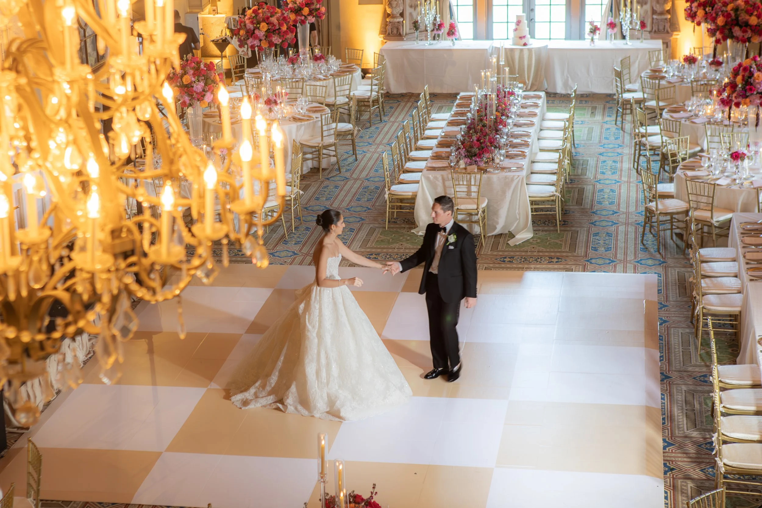 Bride and groom dancing in a wedding reception hall decorated with pink and red flowers, golden chairs, and a large chandelier.